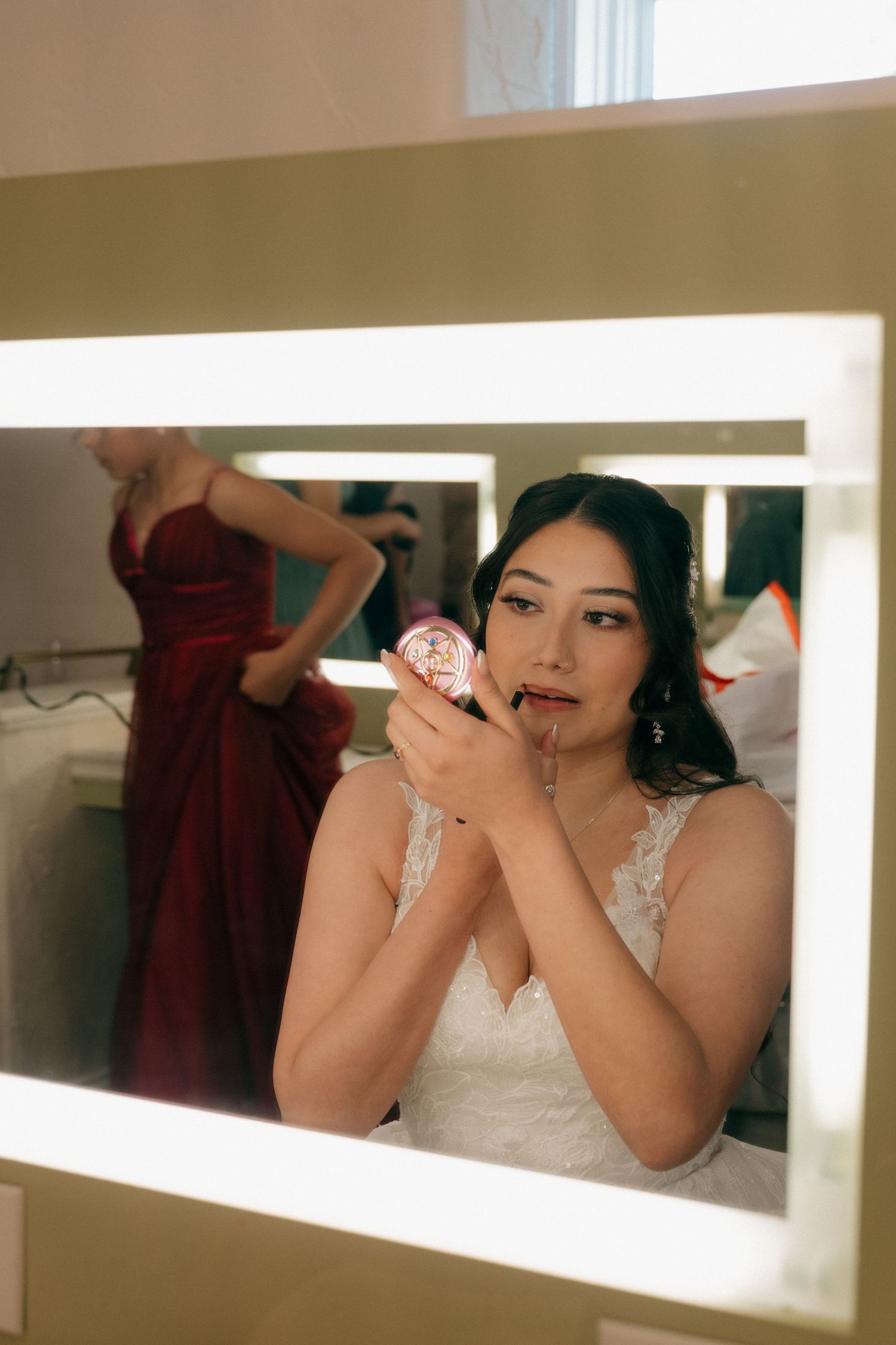 Bride applies lipstick using a compact mirror with lit vanity lights reflecting a bridesmaid in a red gown.
