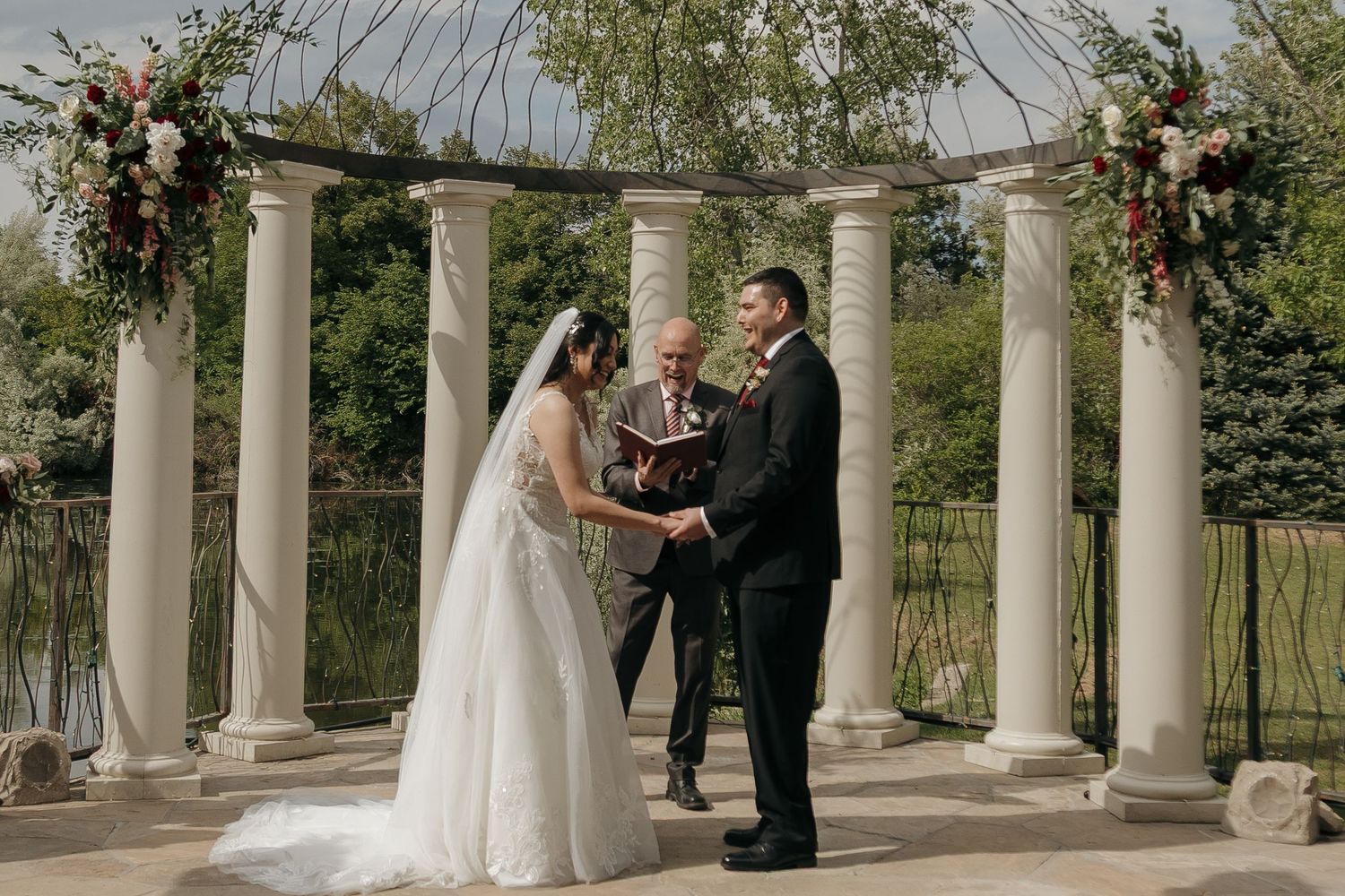 Bride and groom exchange vows under a white columned pergola decorated with floral arrangements outdoors.