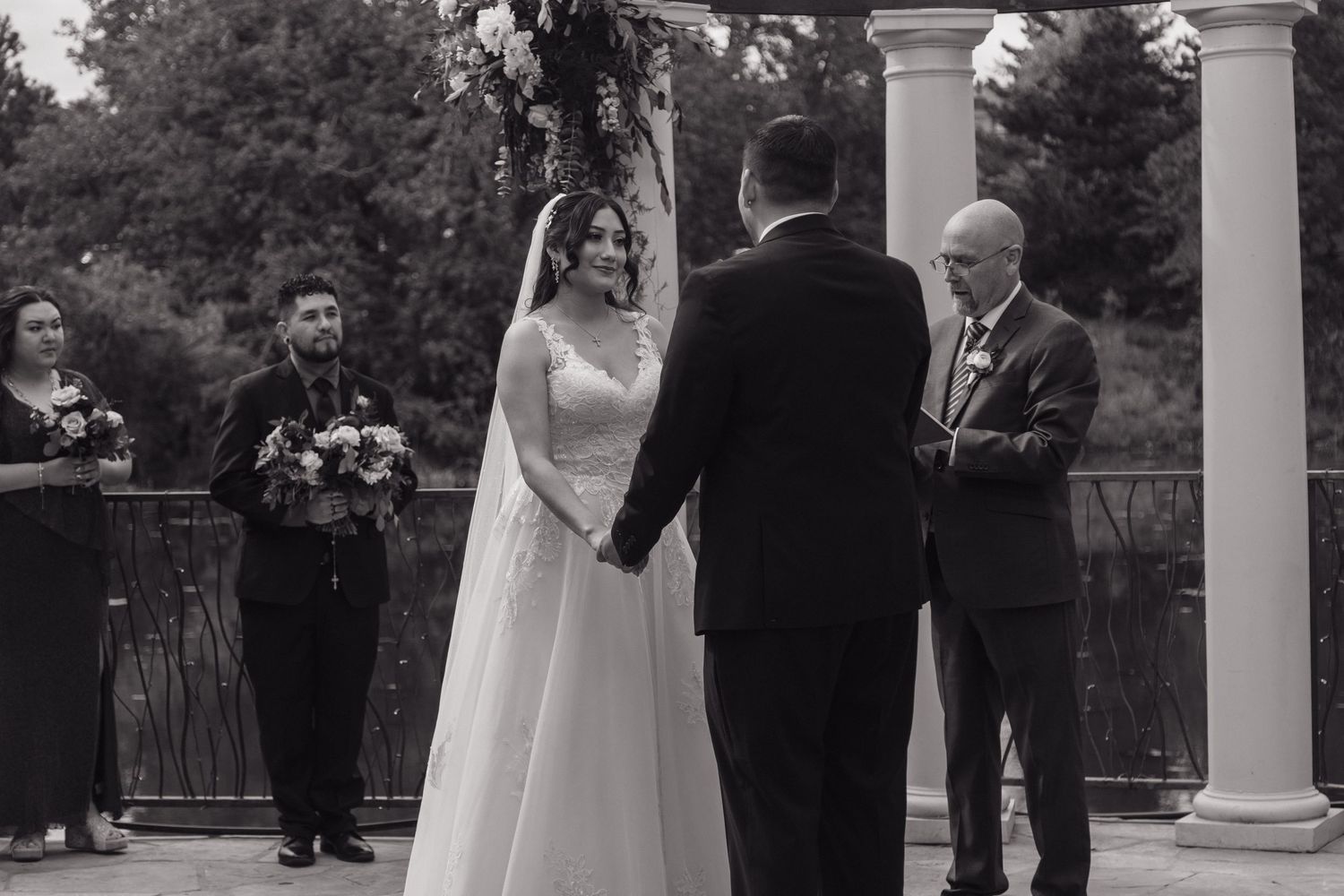 Black and white photo of bride and groom holding hands at outdoor wedding altar with floral arch backdrop.