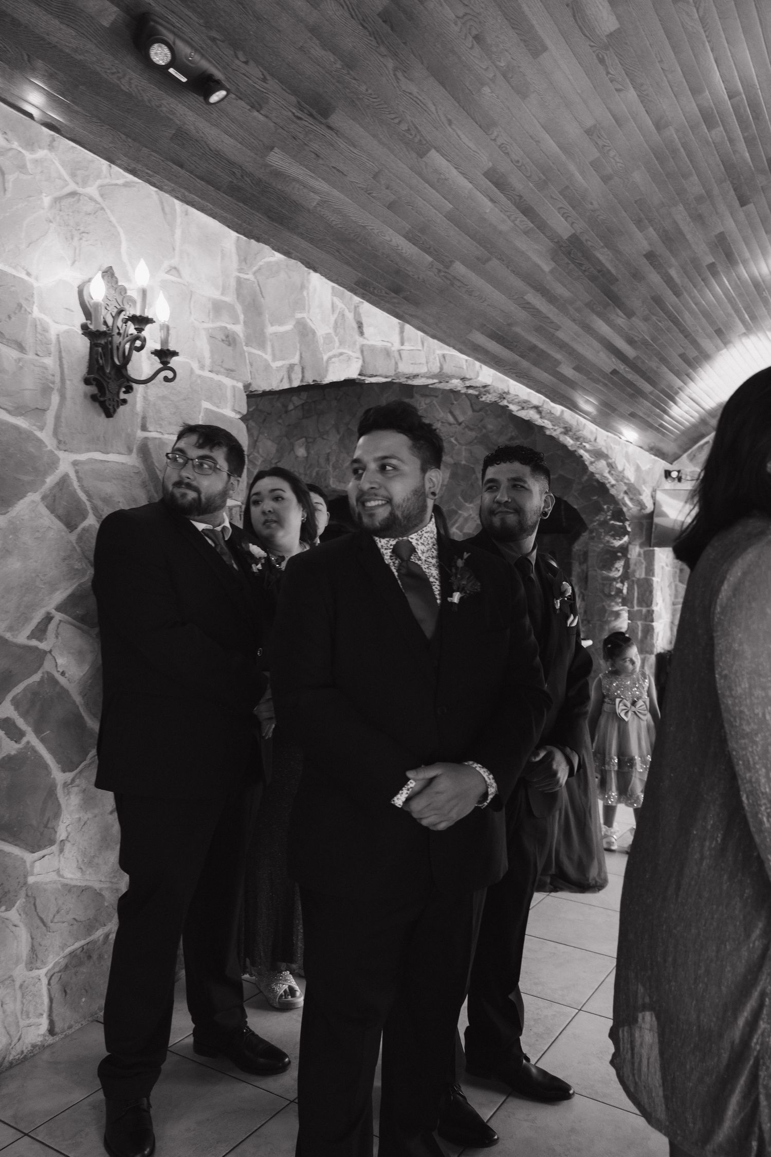 Groomsmen in black suits stand together in a stone-walled venue corridor during a wedding ceremony.