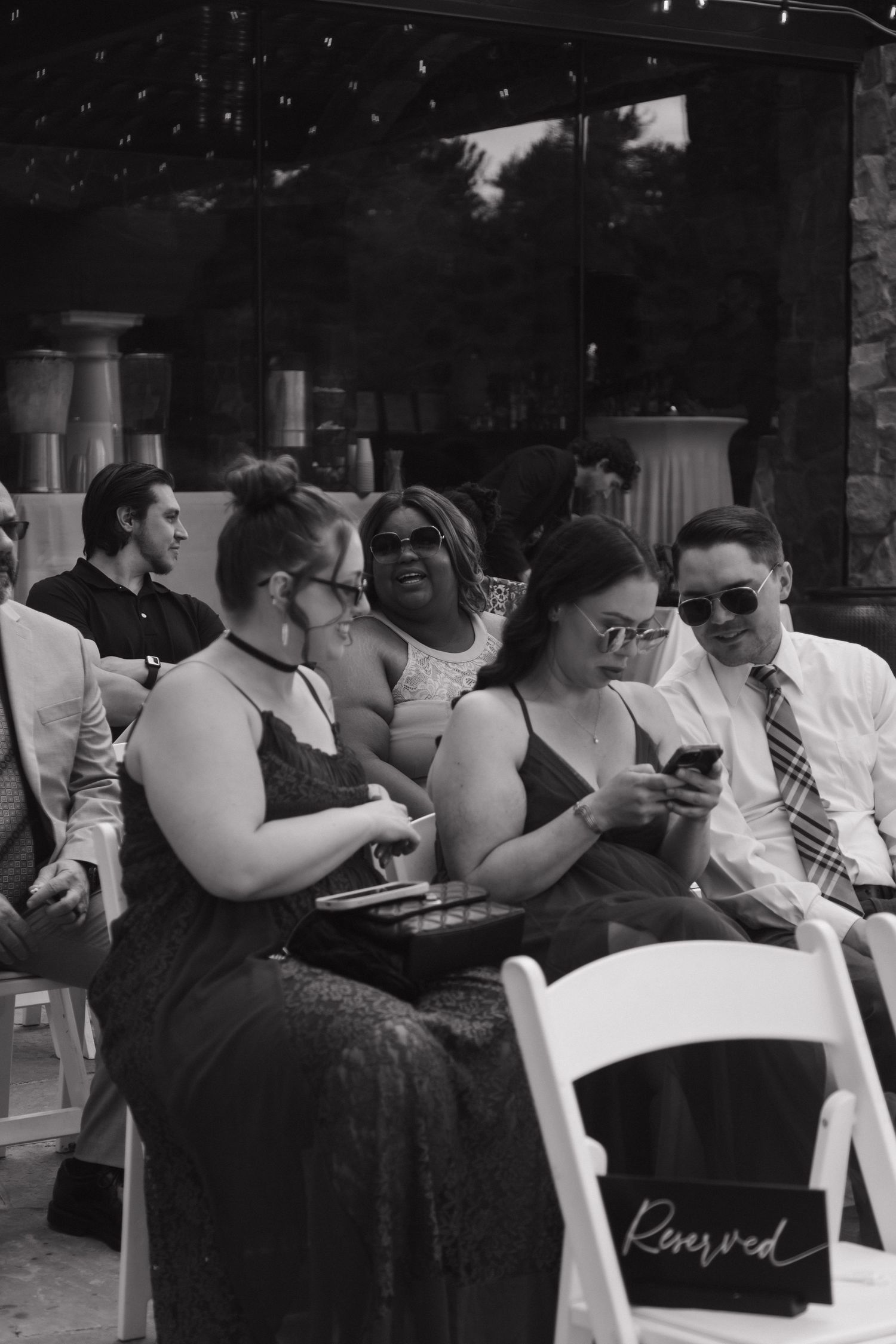 Wedding guests smile and look at phones while seated in white chairs at an outdoor ceremony.