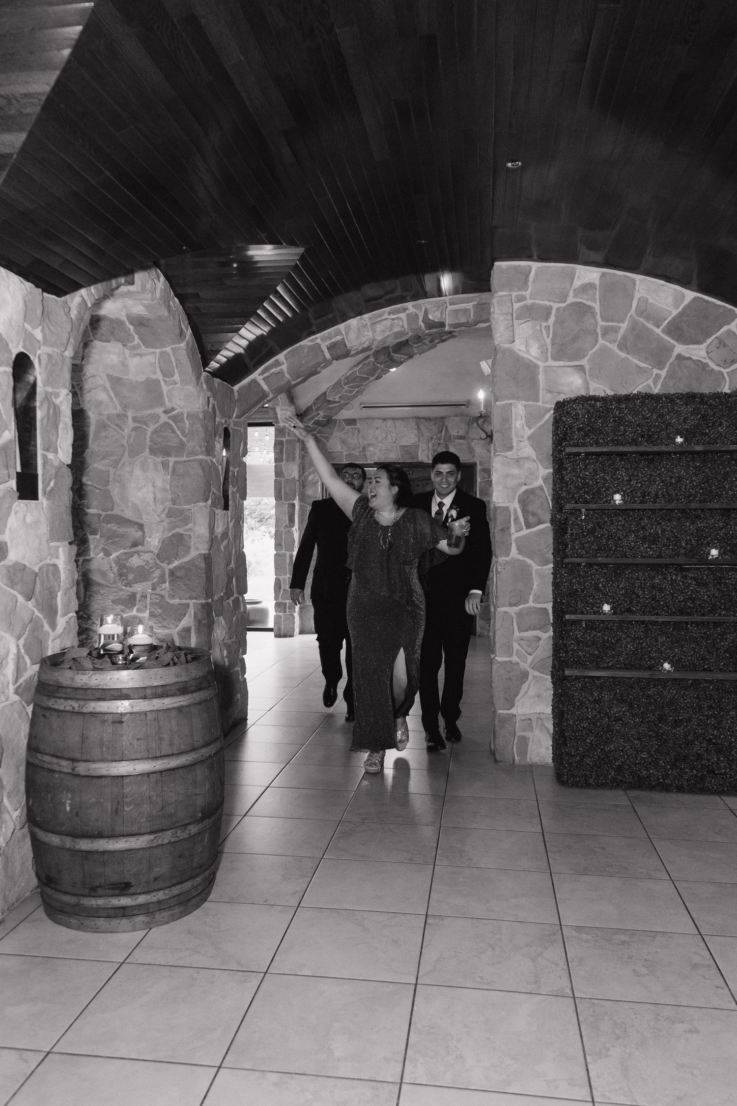 Wedding guests walk through a stone archway corridor with barrel decor at a rustic venue.
