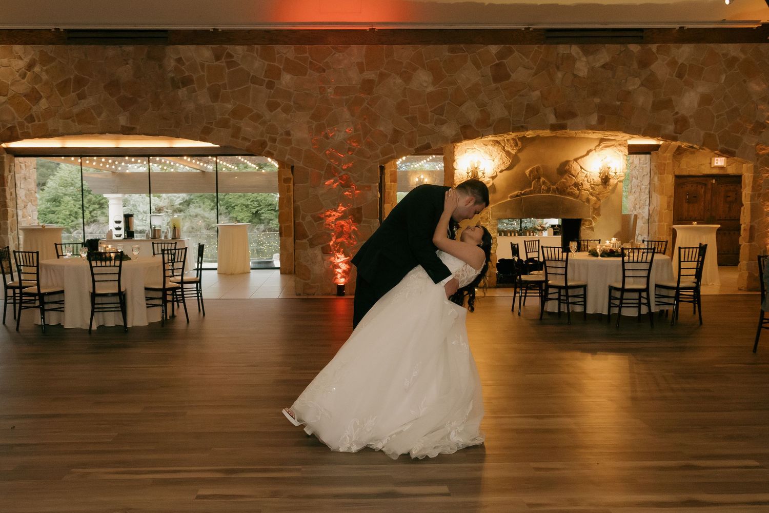 Groom dips bride during their first dance in a warmly lit rustic stone reception hall.