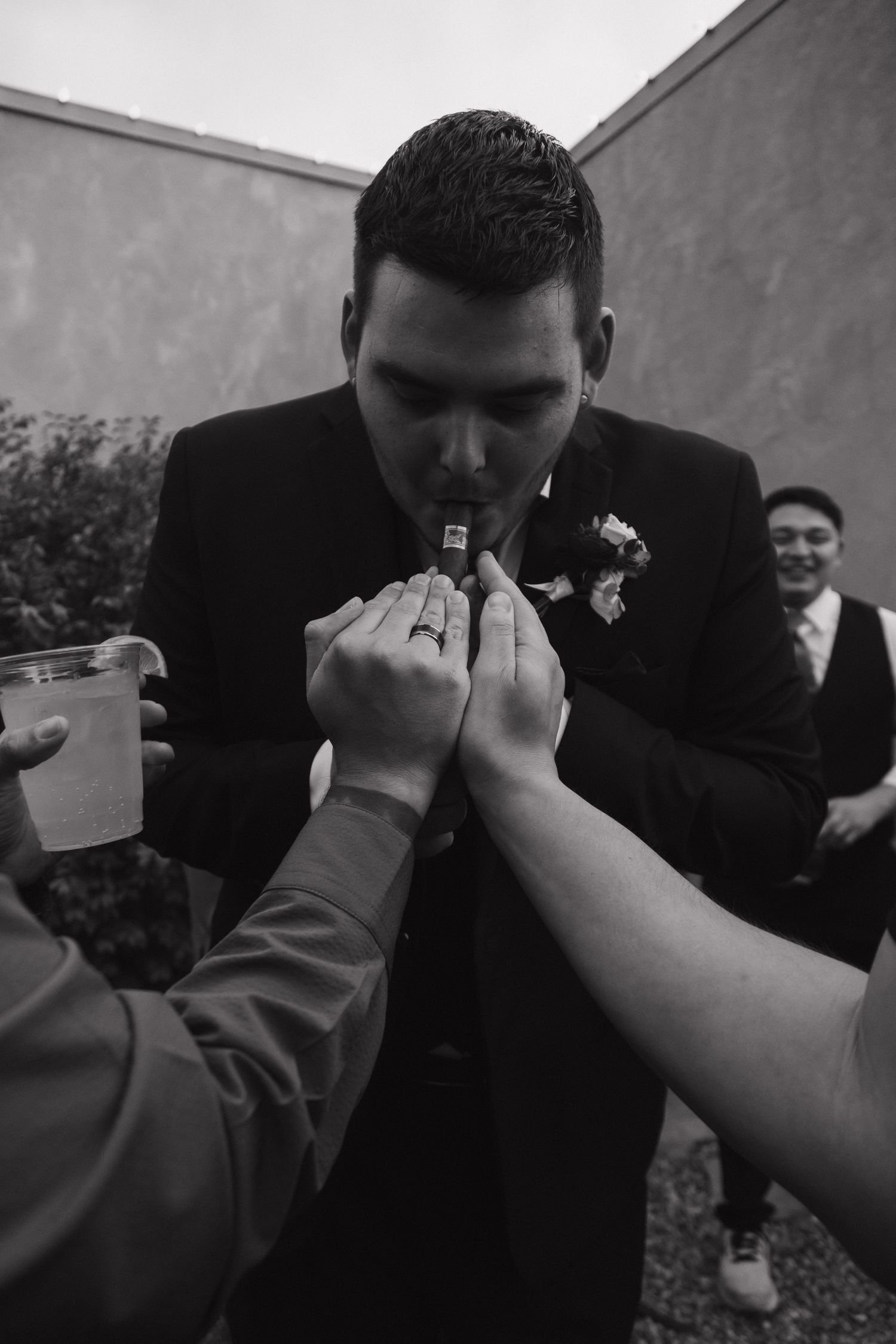 Groom in black suit lights a cigar assisted by guests at an outdoor wedding celebration.