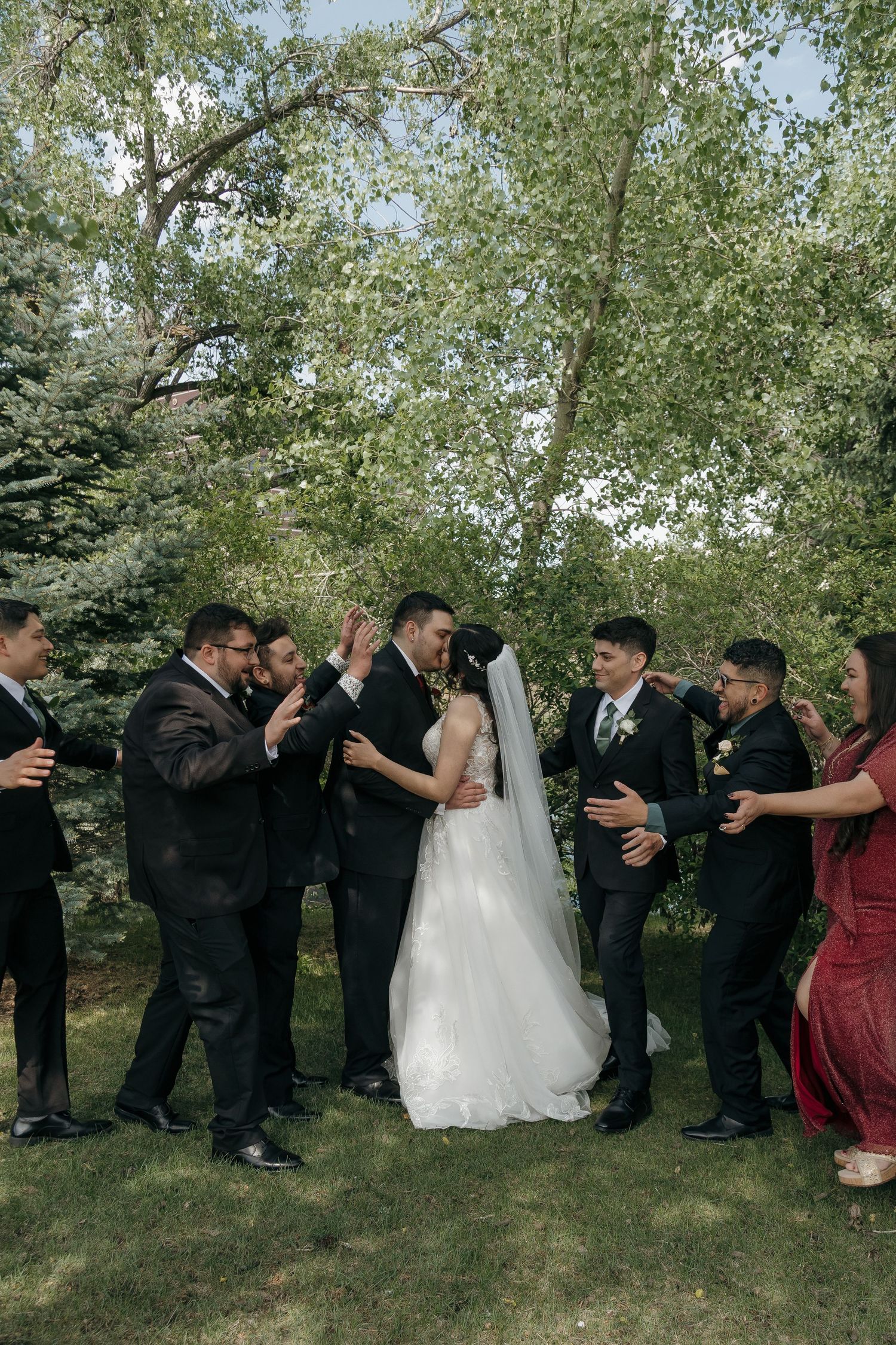 Bride and groom kissing surrounded by cheering wedding party in black suits and colorful dresses outdoors.