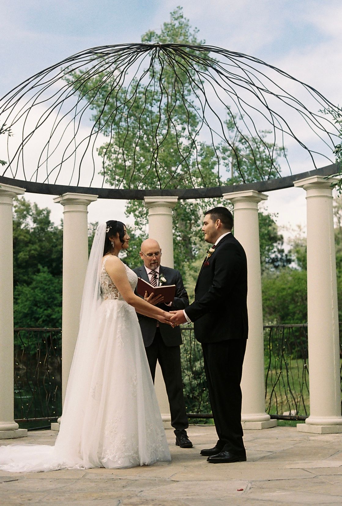 Bride and groom exchange vows with officiant under elegant iron dome gazebo with white columns.