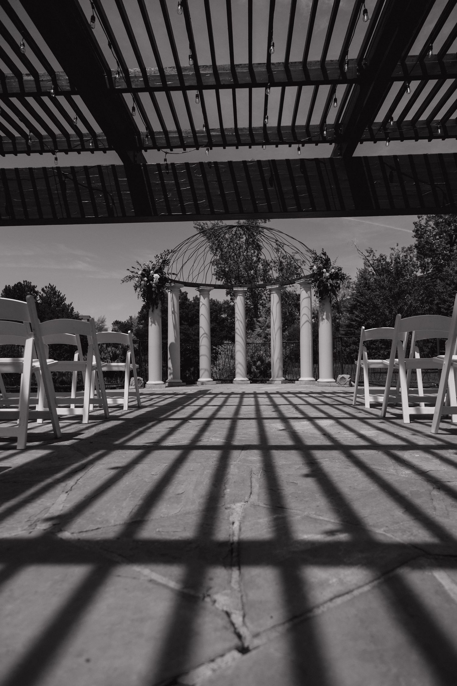 Black and white photo of elegant outdoor wedding ceremony venue with white chairs and floral arch.