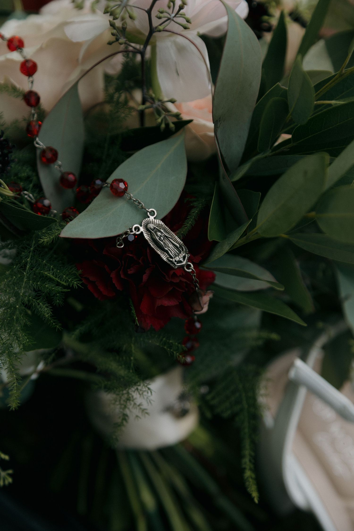 A bridal bouquet with red roses and greenery adorned with a silver rosary and religious medal.