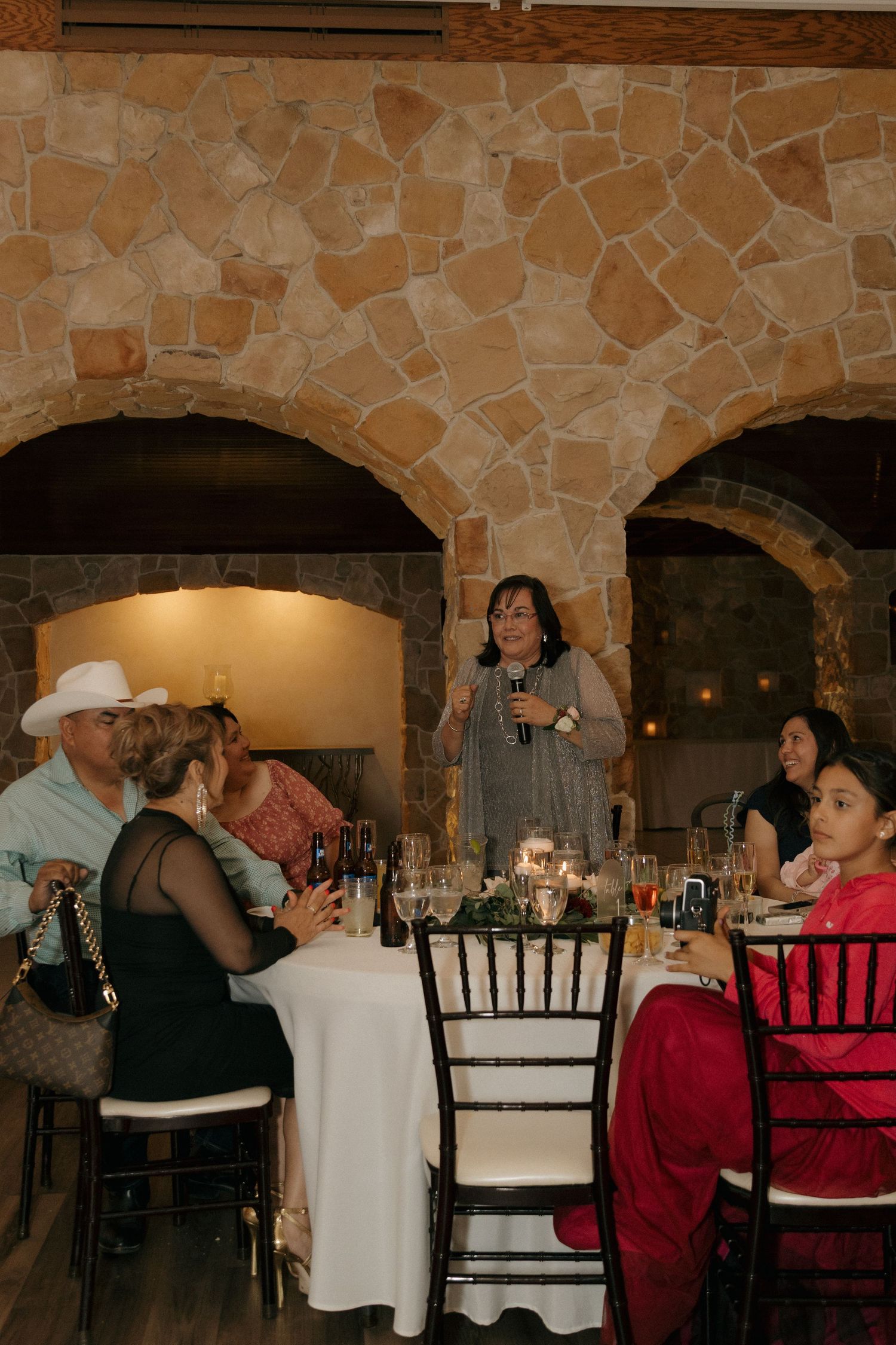 Wedding guest giving a speech with microphone at a candlelit table in a stone venue.