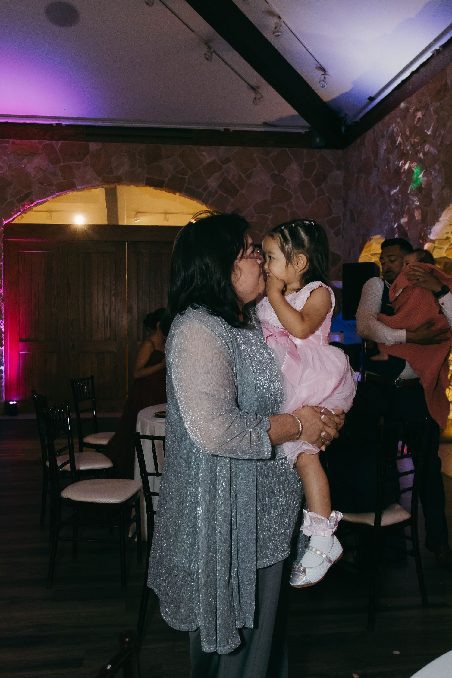 Grandmother shares a tender kiss with young flower girl in pink dress at wedding reception.