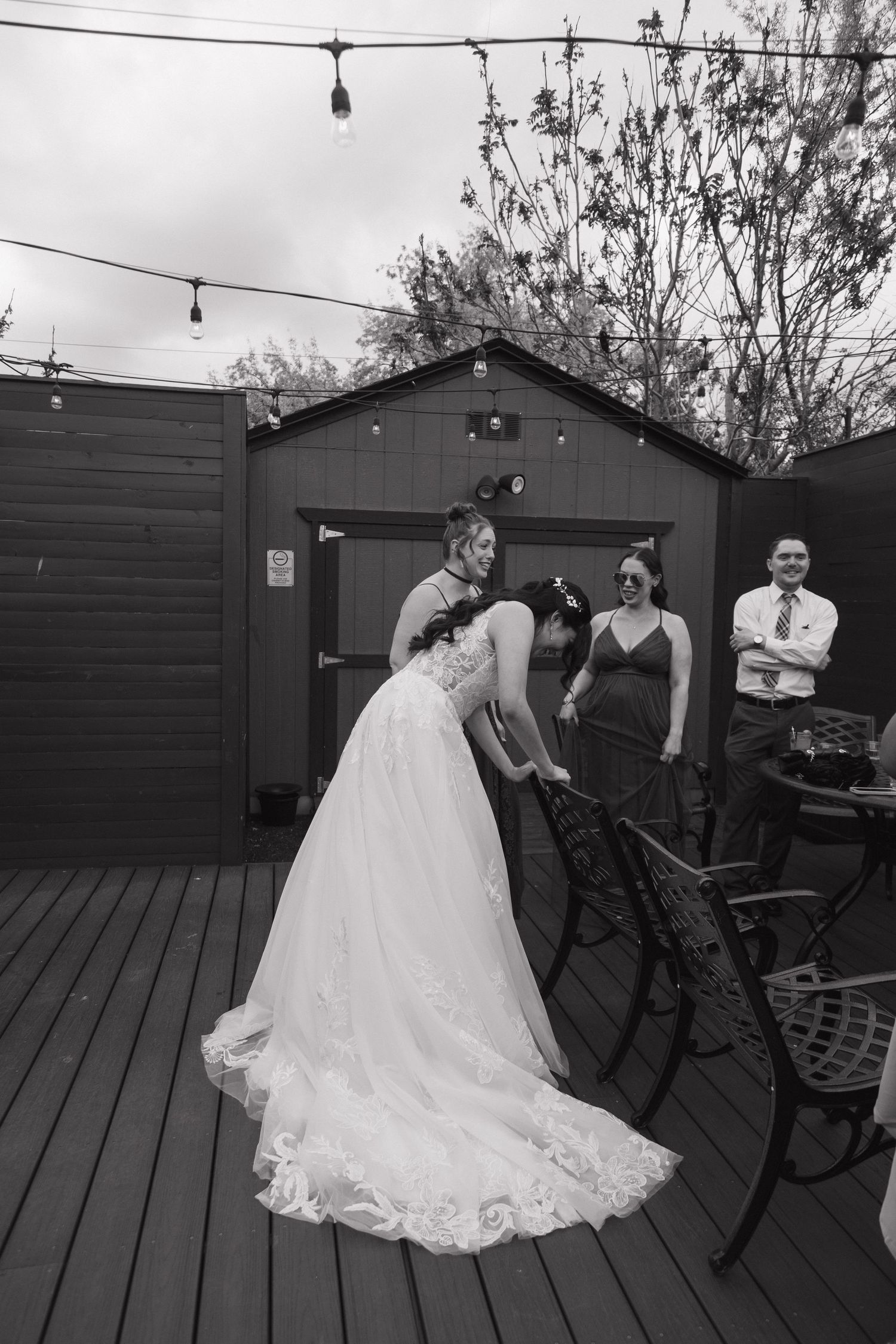 Black and white photo of bride in lace gown being assisted by wedding party on outdoor wooden deck.
