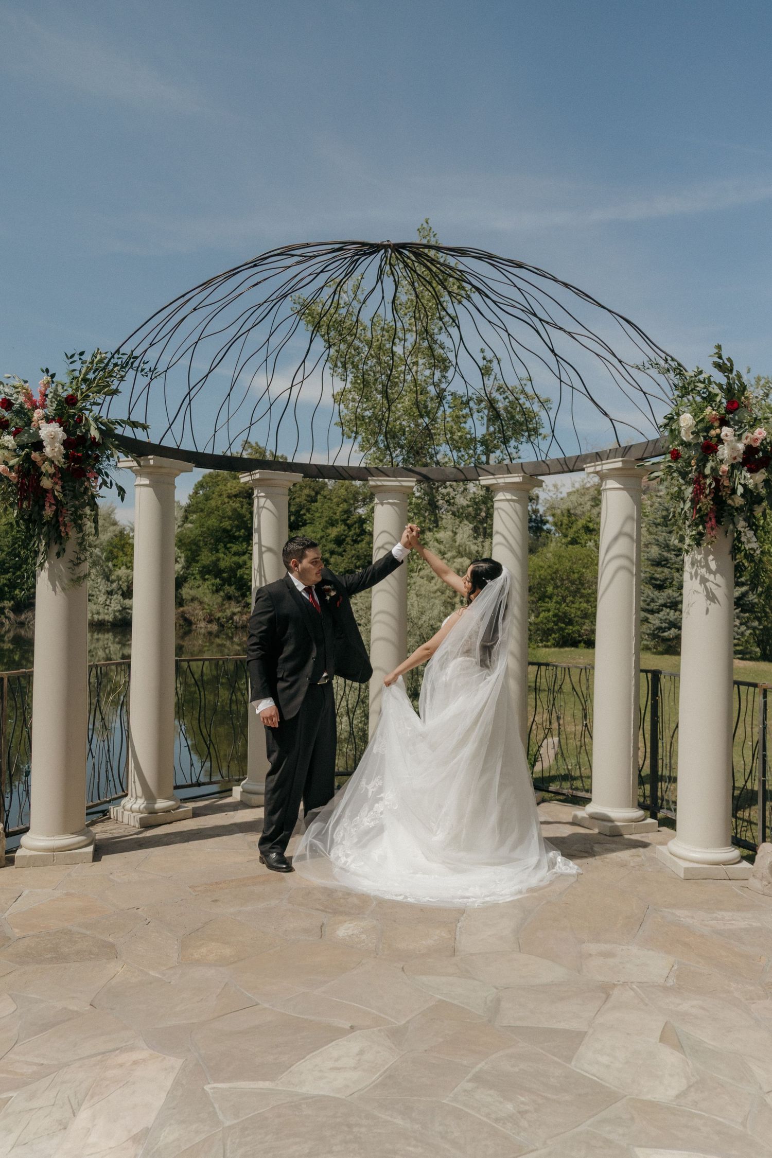 Bride and groom dance joyfully under an elegant iron gazebo with floral accents beside a tranquil lake.