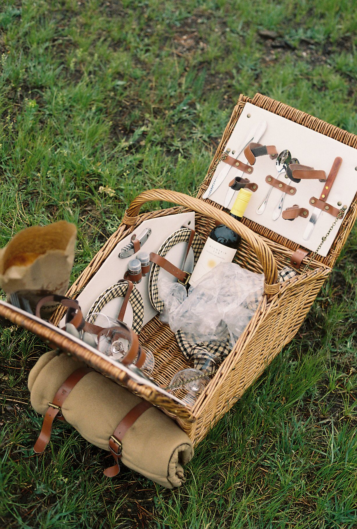 Open wicker picnic basket filled with supplies and utensils sitting on green grass outdoors.