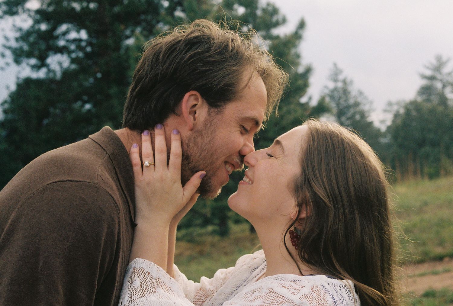 Engaged couple sharing a romantic kiss outdoors with pine trees in the soft background.