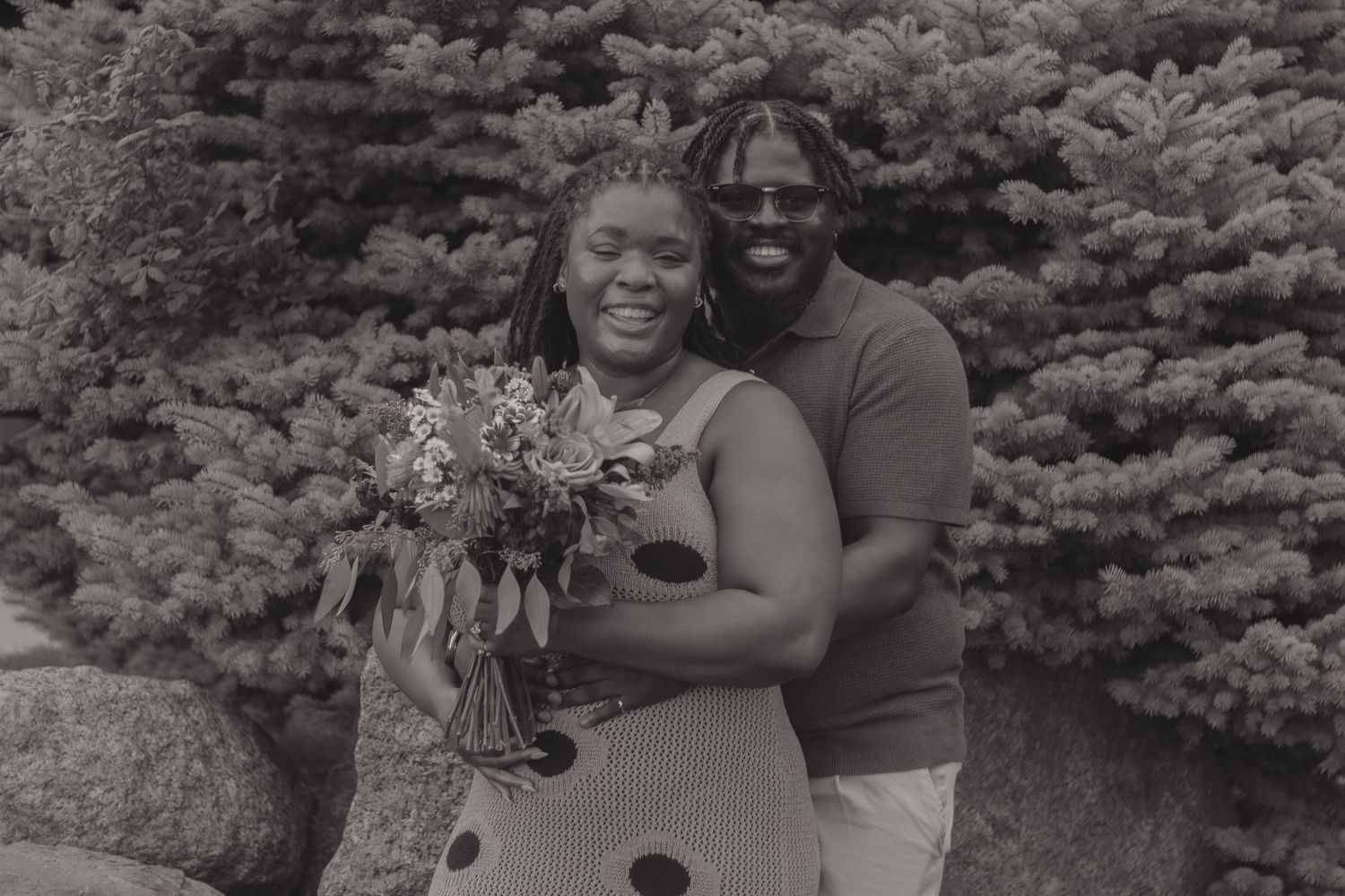 A joyful couple embraces while holding a bouquet against an evergreen backdrop.