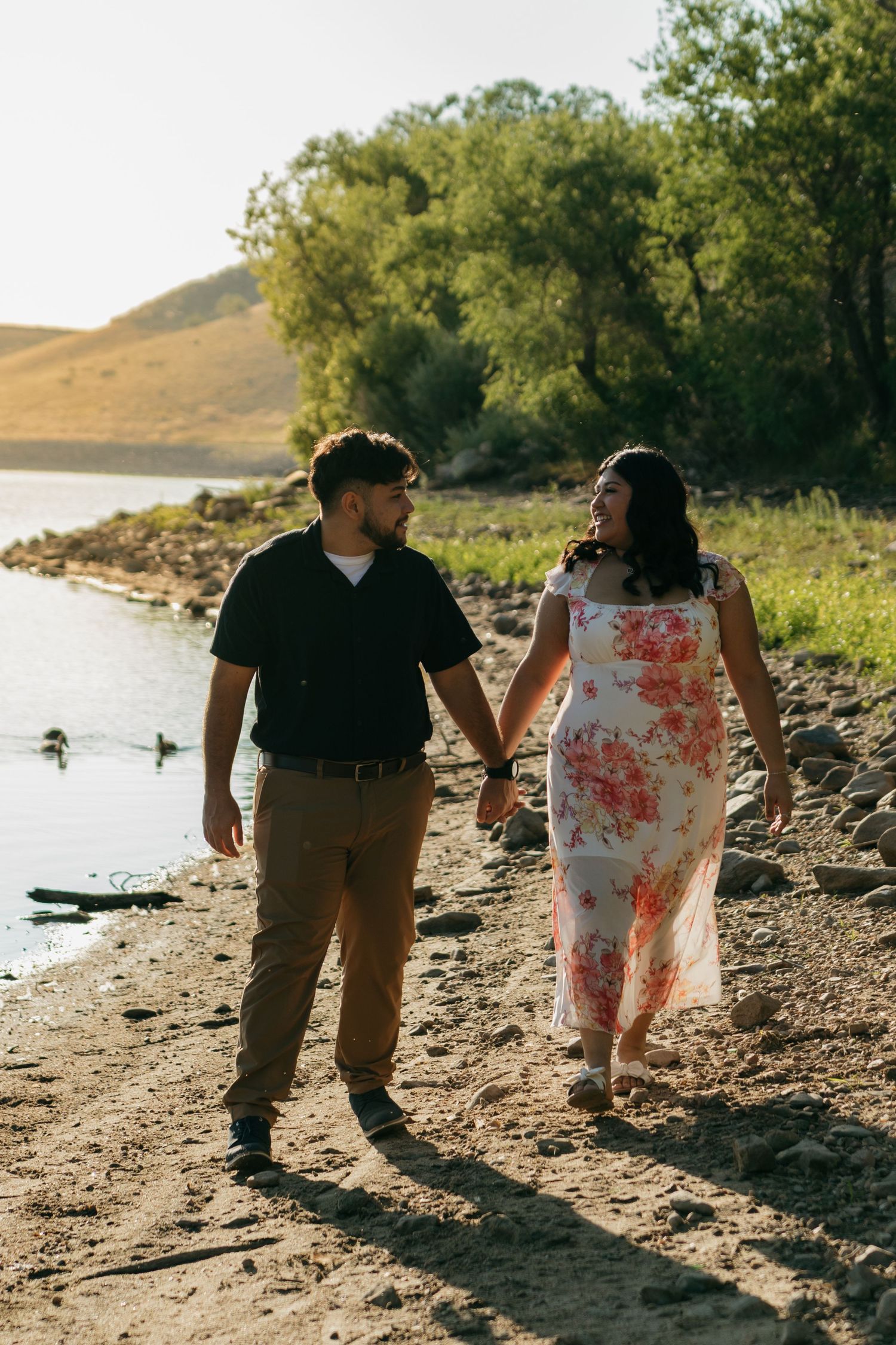 A couple walks hand in hand along a lakeside trail during golden hour, with her in a floral dress and him in casual wear.