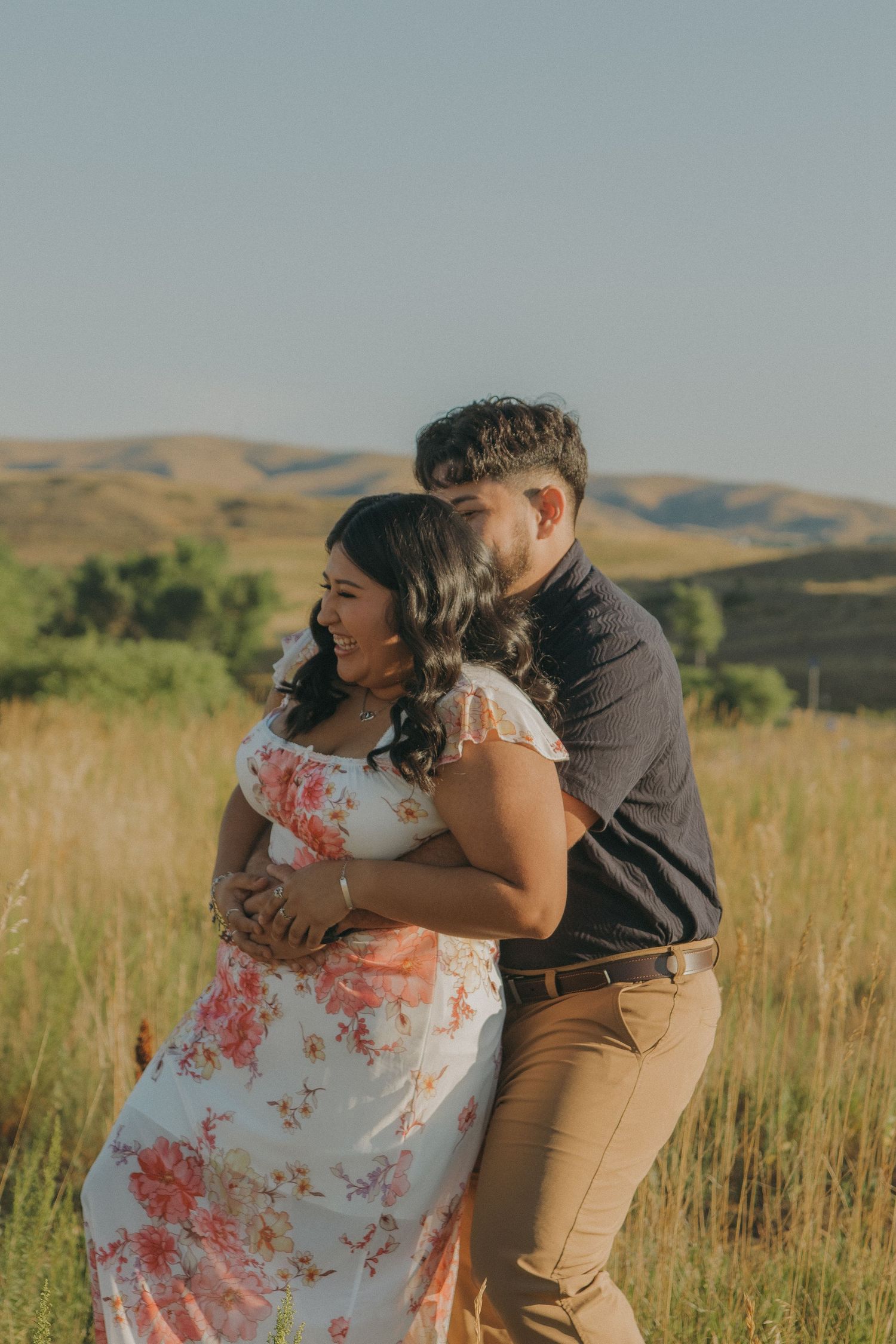 A couple shares an intimate moment in tall grass with rolling hills in the background.