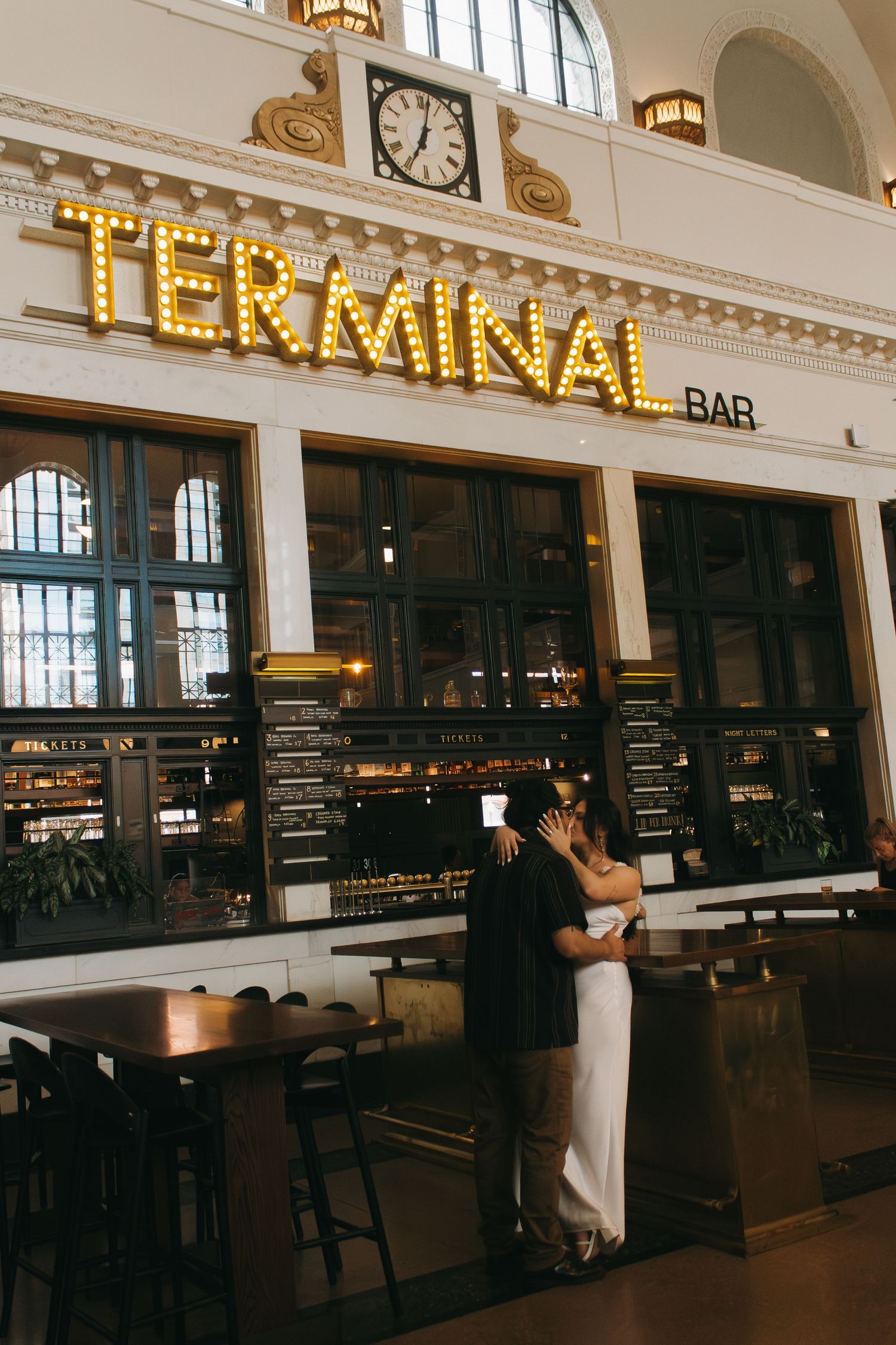 Couple embracing at Terminal Bar under illuminated vintage marquee sign in historic train station interior.