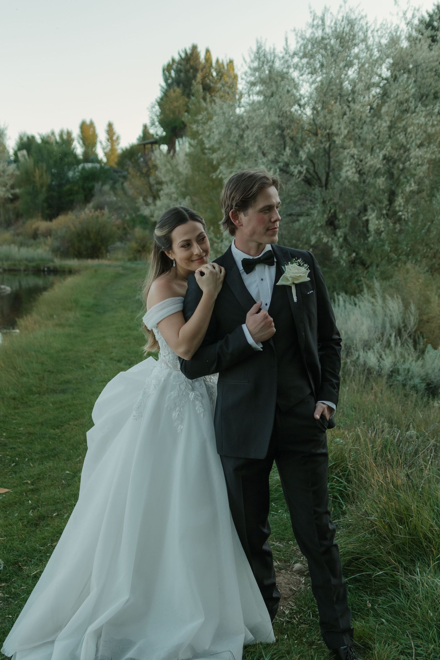 A couple in wedding attire poses together outdoors on a grassy path with willow trees in the background at dusk.