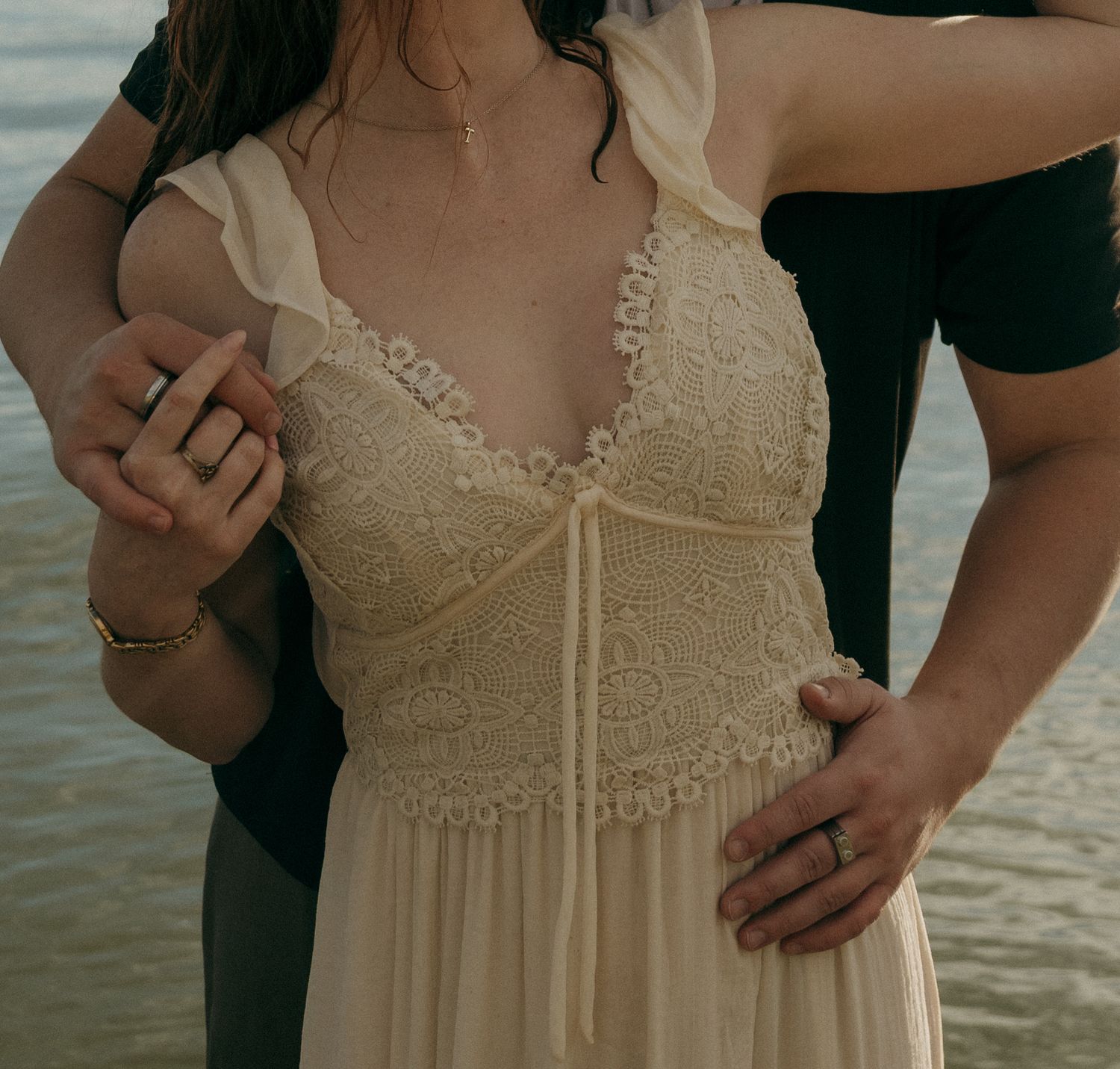 Close-up of woman in cream lace boho dress with man's hands on her waist near water.