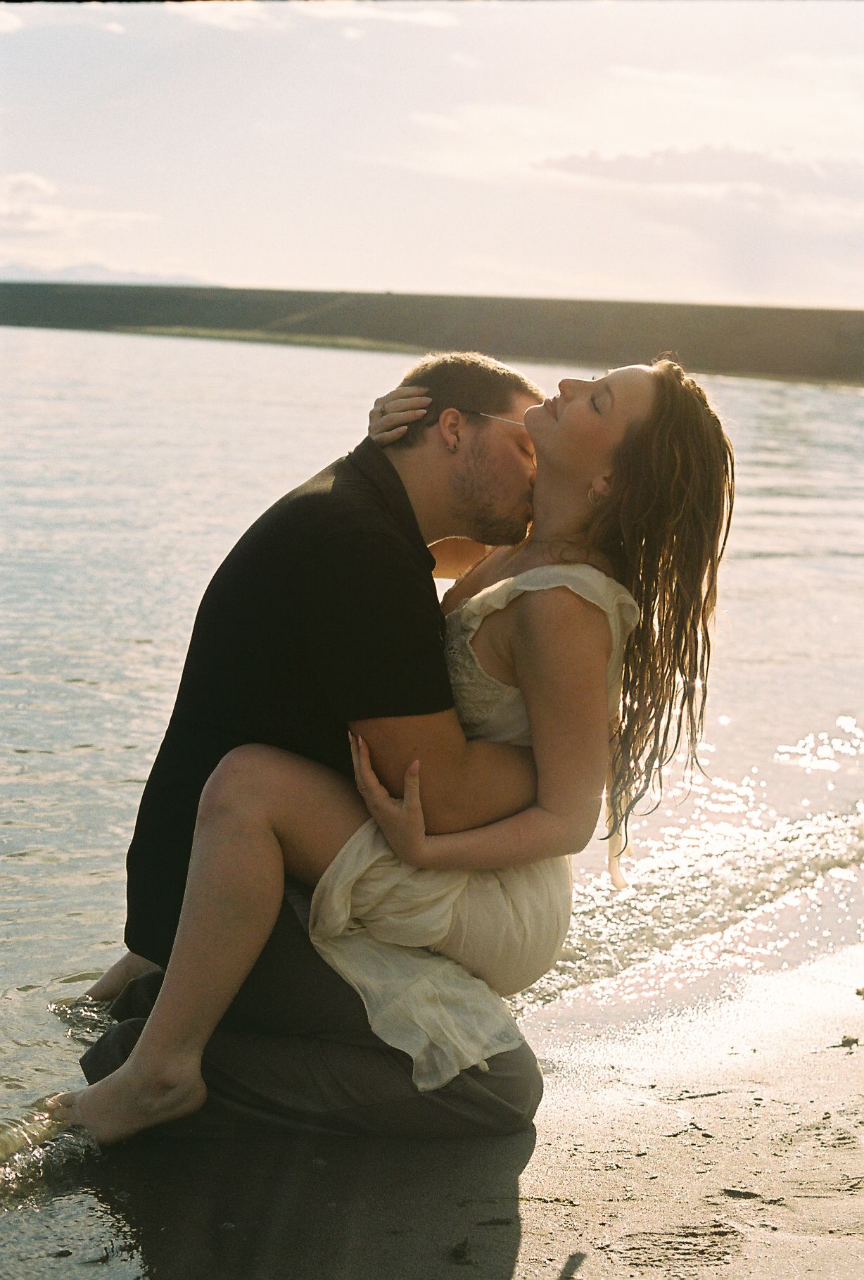 Man carrying woman in white dress while kissing on a sunlit rocky shoreline.