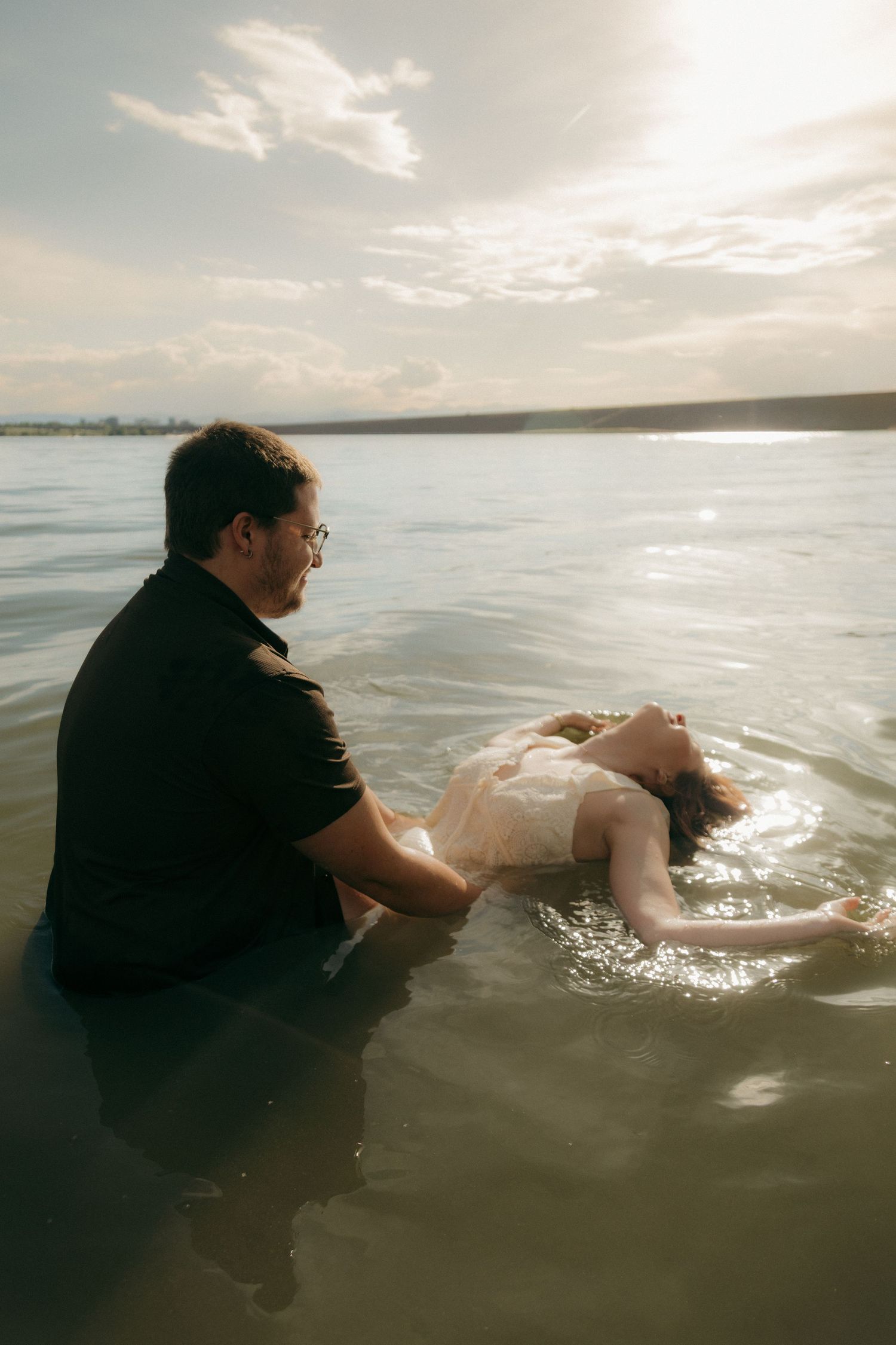 Man sitting in shallow lake water holding woman floating on her back in white dress at sunset.