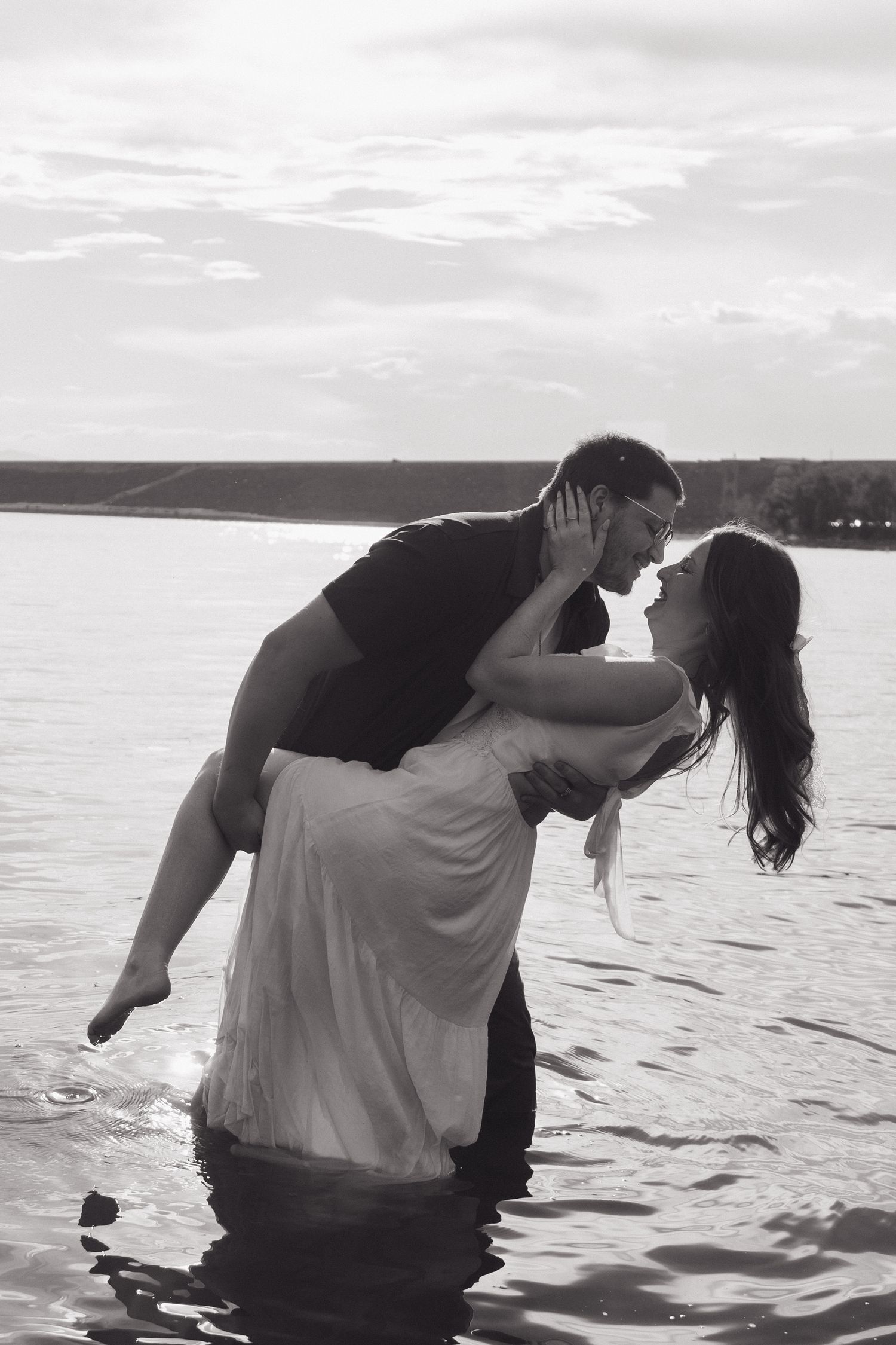 Black and white photo of couple kissing in water, man lifting woman in white wedding dress at sunset.
