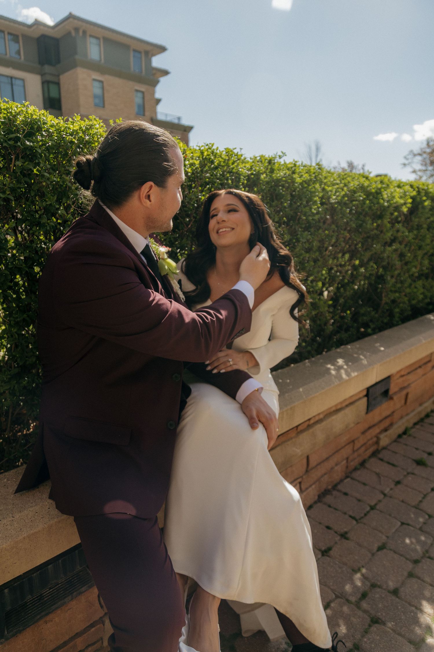 Groom in burgundy suit tenderly touches brides face as she smiles while seated on stone ledge outdoors.