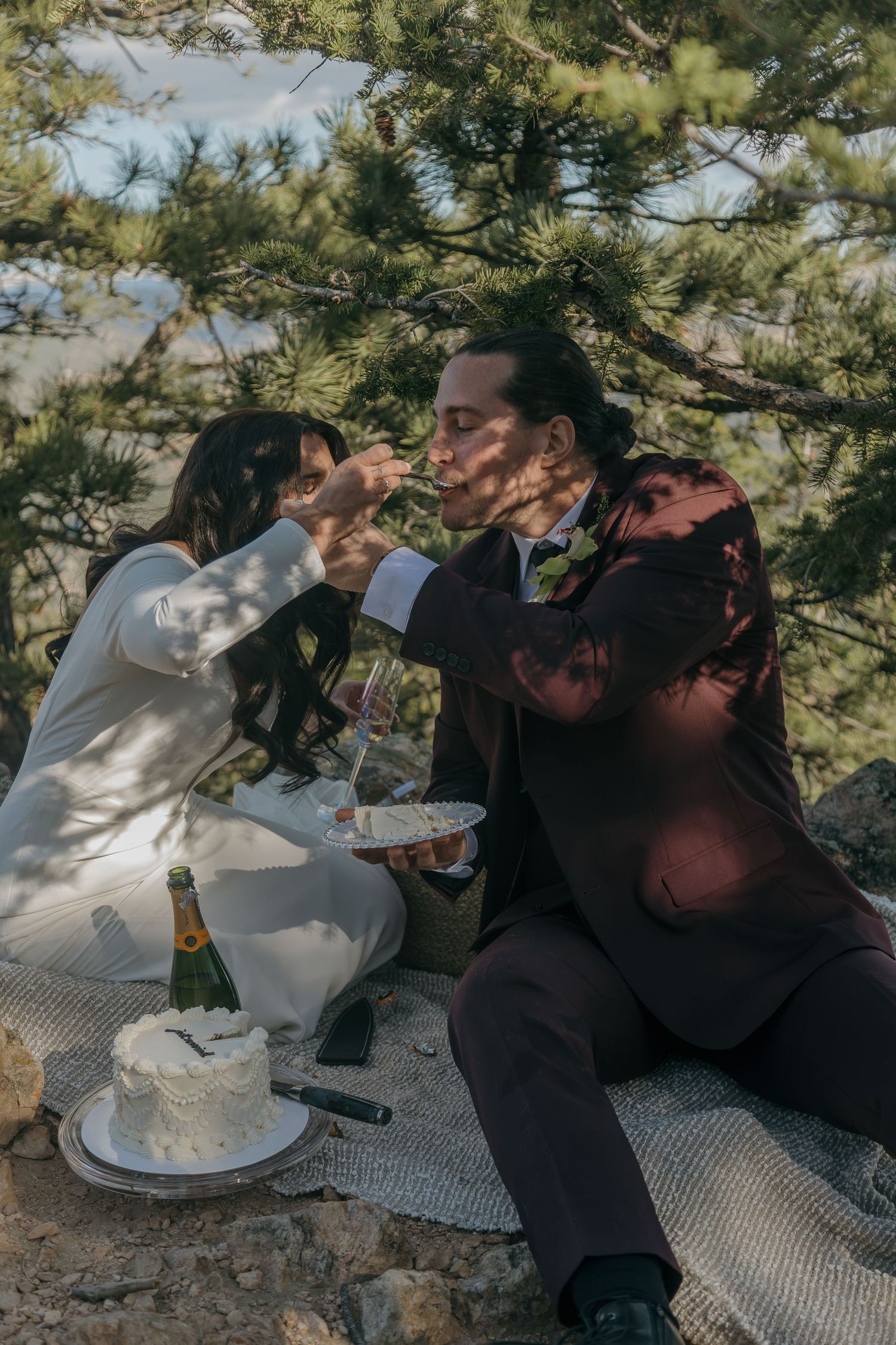 Bride feeding groom wedding cake while sitting outdoors under pine trees with Veuve Clicquot champagne.