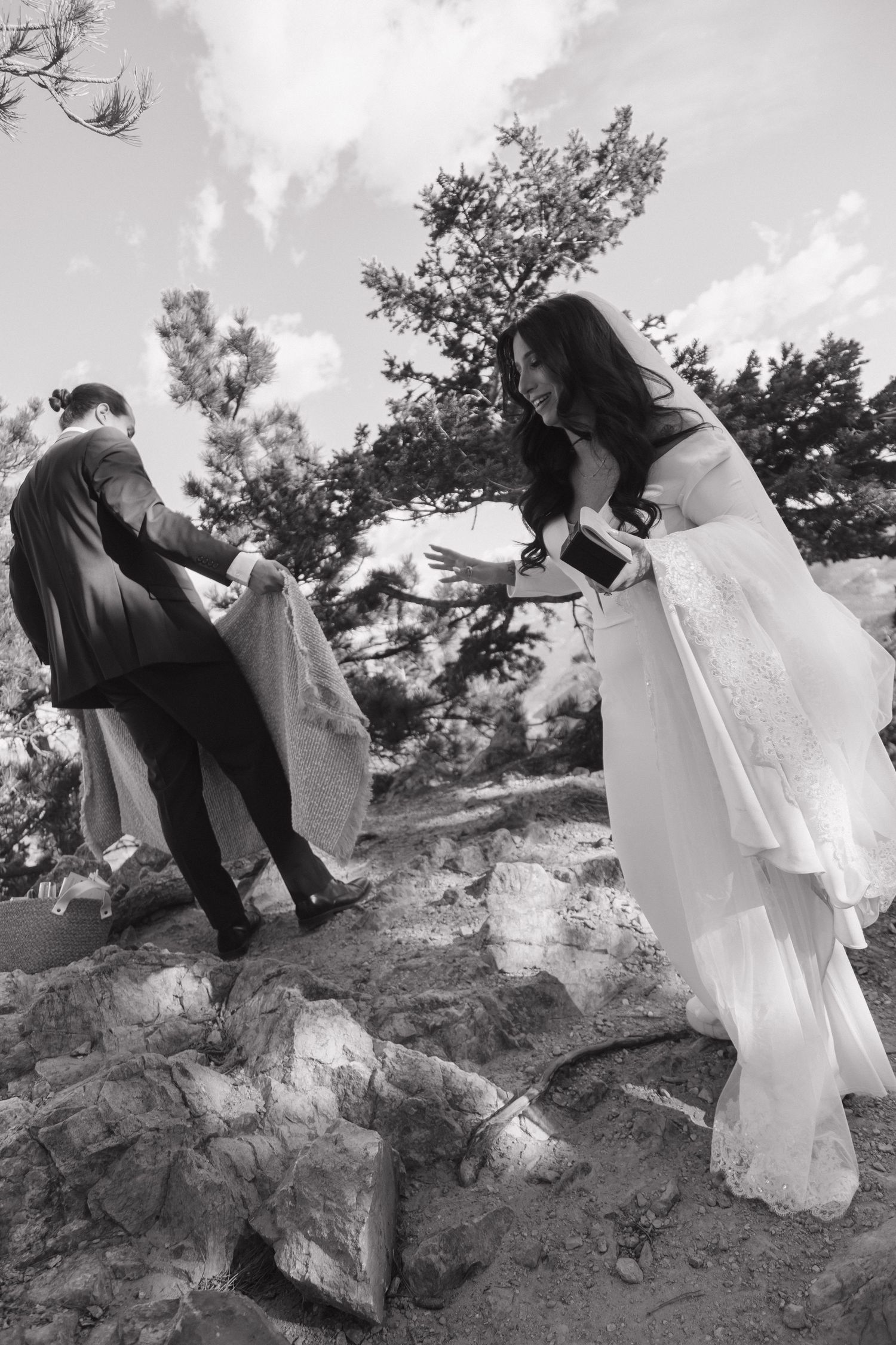 Black and white photo of bride and groom helping each other over rocks at outdoor wedding.
