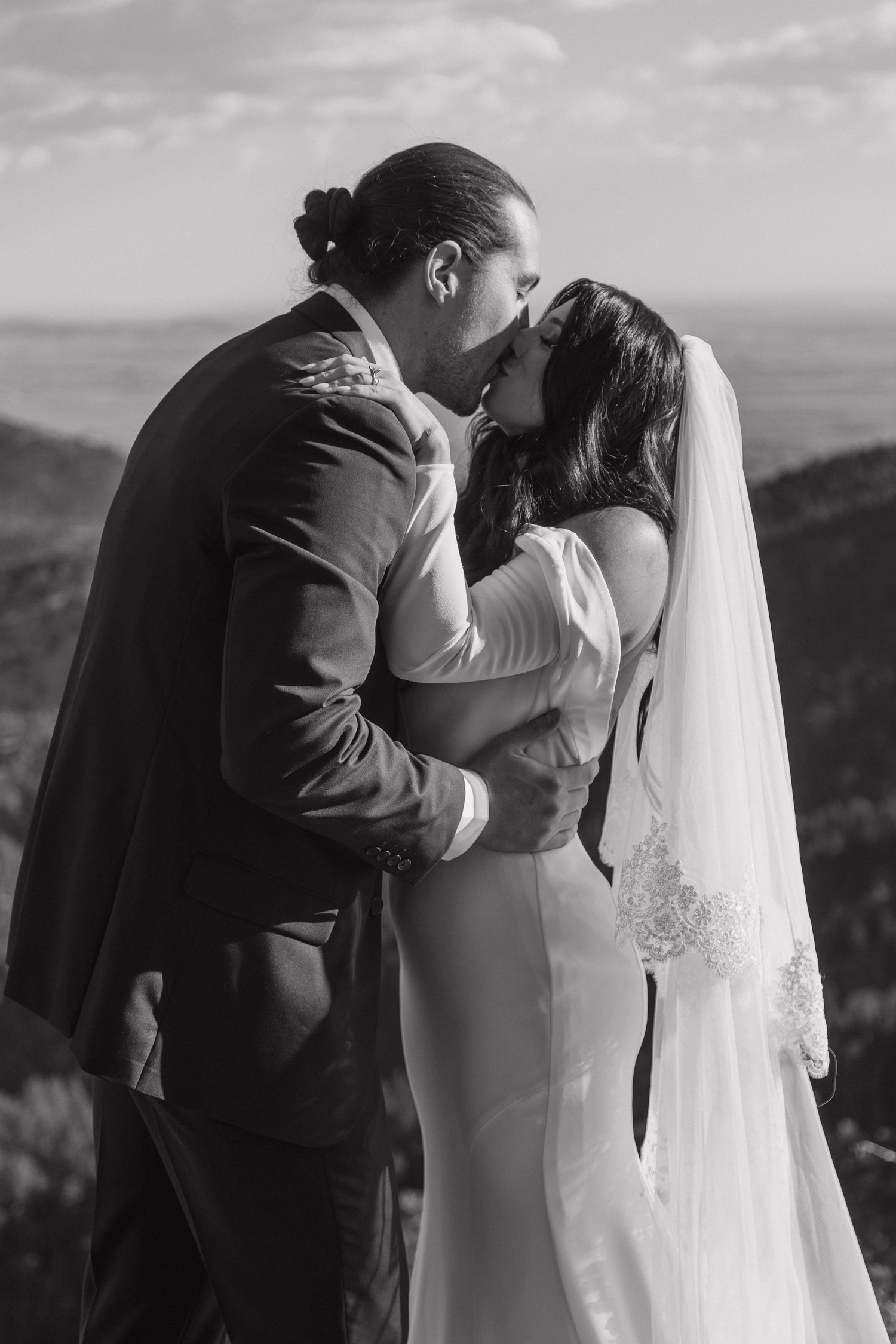 Black and white photo of bride and groom sharing first kiss on scenic mountain overlook.