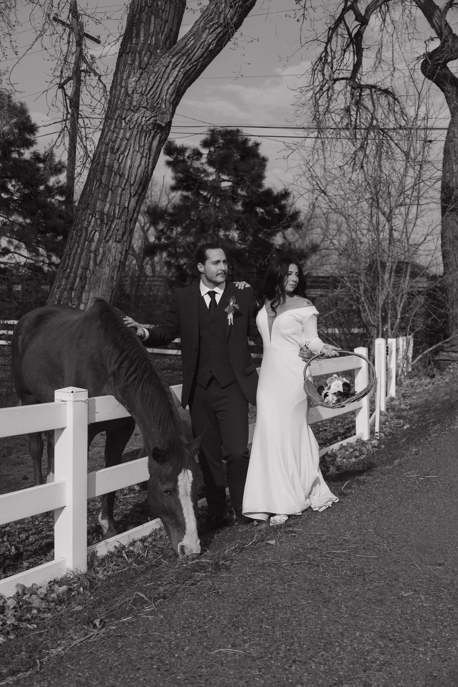 Black and white photo of bride and groom walking beside a horse along a white fence.