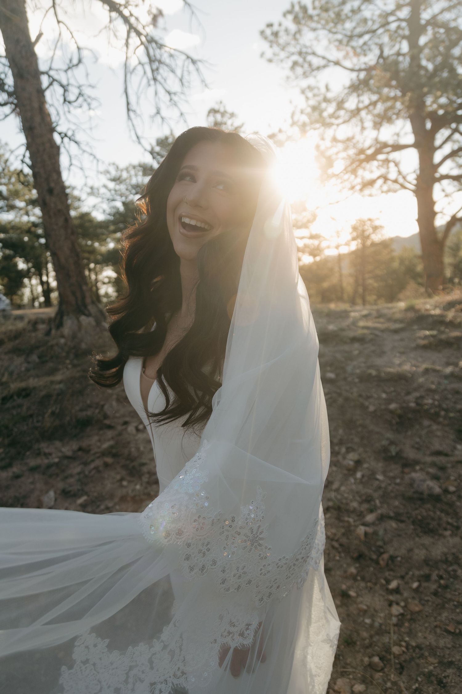 Joyful bride in white gown and embroidered veil laughing in sunlit pine forest during golden hour.
