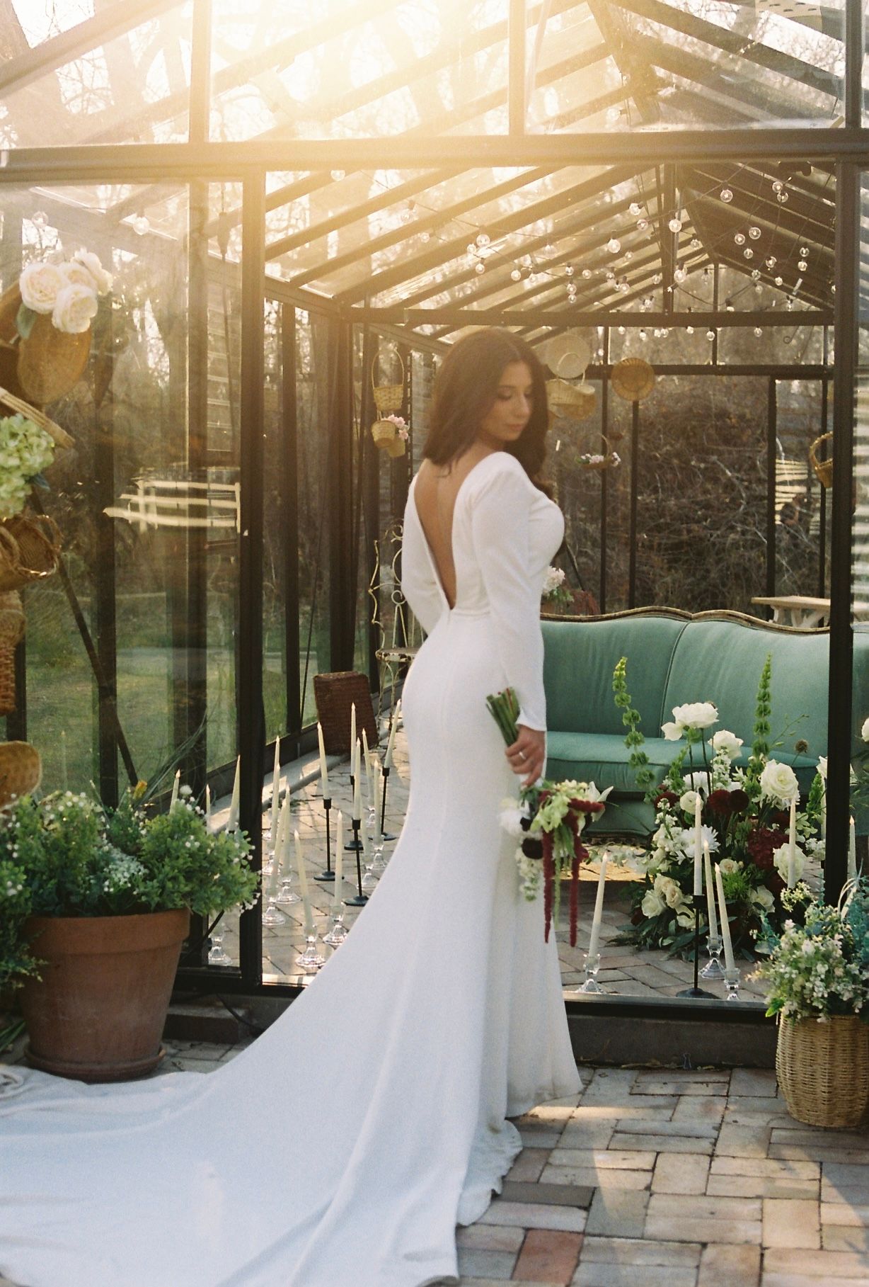 Bride in elegant backless white gown posing inside a sunlit greenhouse surrounded by candles and florals.