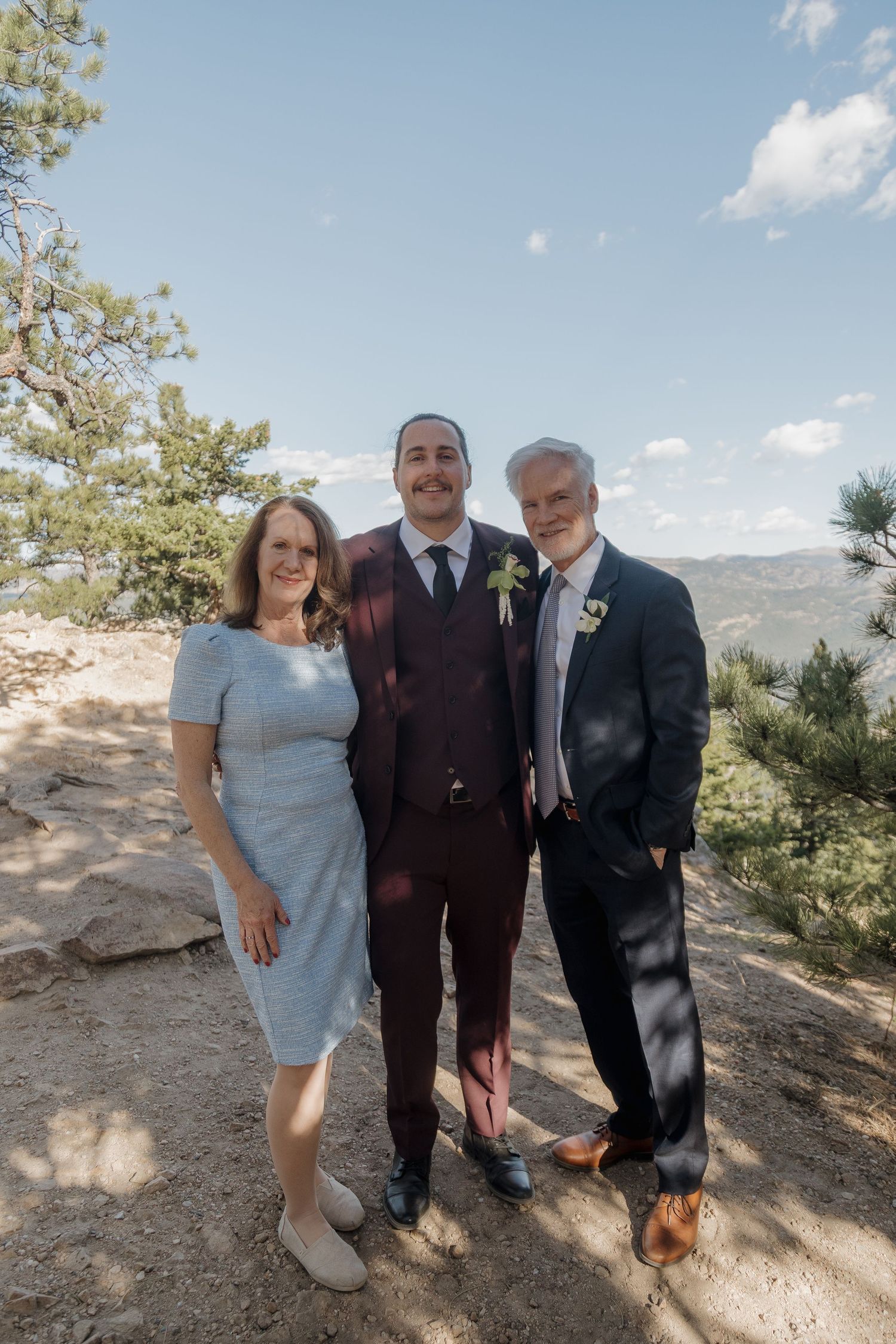 Groom in burgundy suit poses with parents outdoors on a rocky mountain landscape on wedding day.