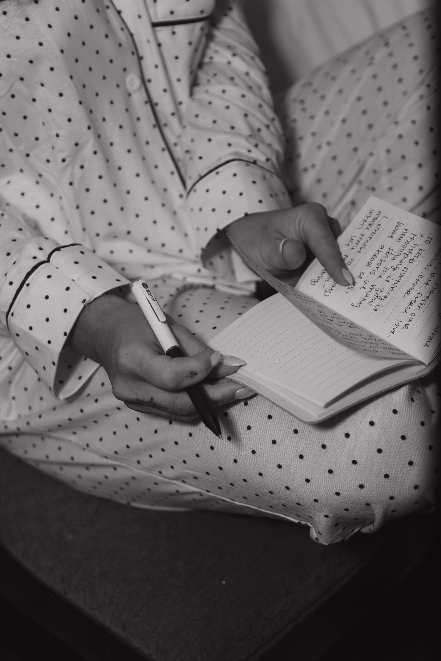 Black and white close-up of bride in polka dot pajamas writing wedding vows in notebook.
