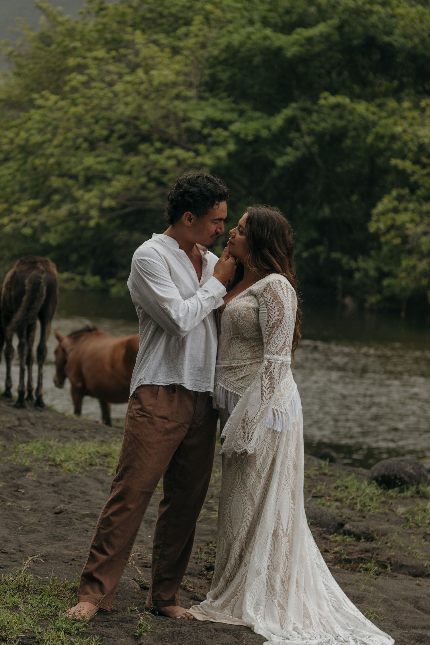 Bride in lace gown and groom in white shirt share an intimate moment with horses by a misty river.