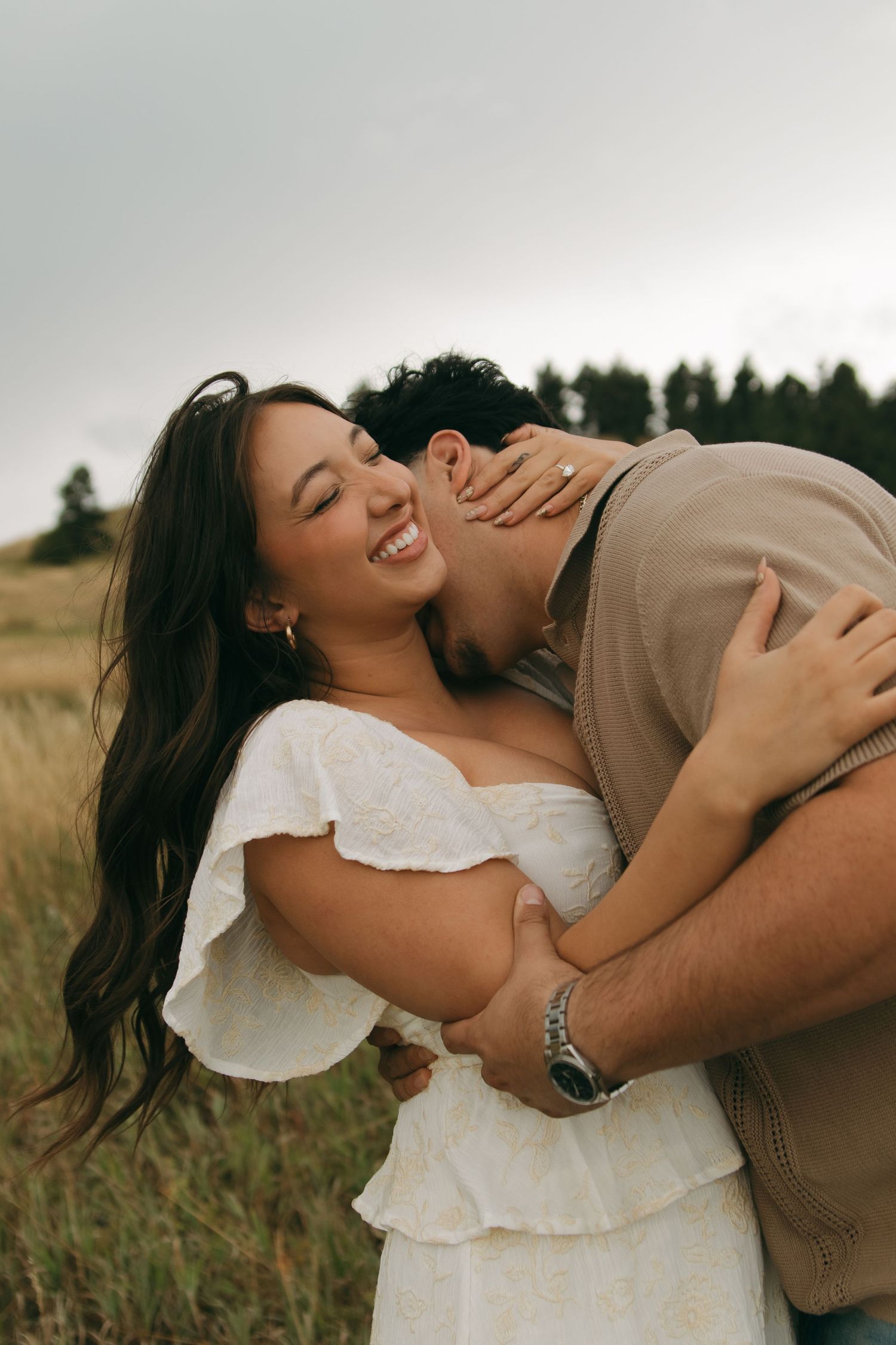 Bride in white dress laughs joyfully as groom kisses her cheek in a golden Colorado meadow.