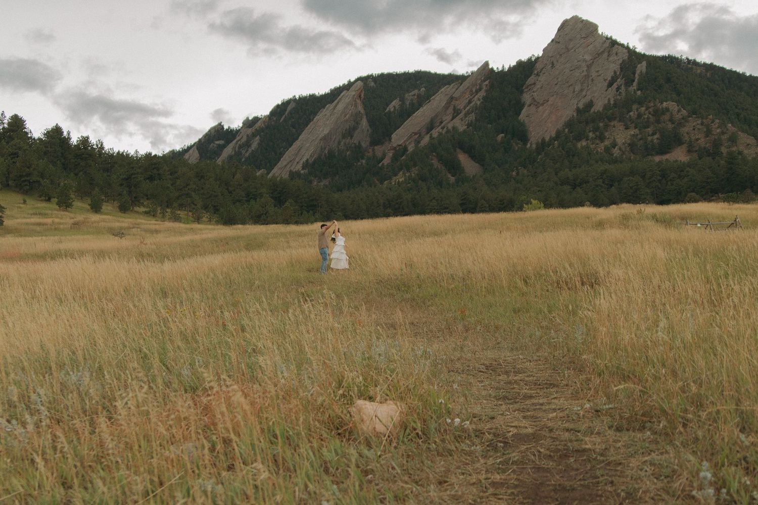 Couple standing together in vast golden meadow with dramatic rocky Flatirons mountain backdrop.