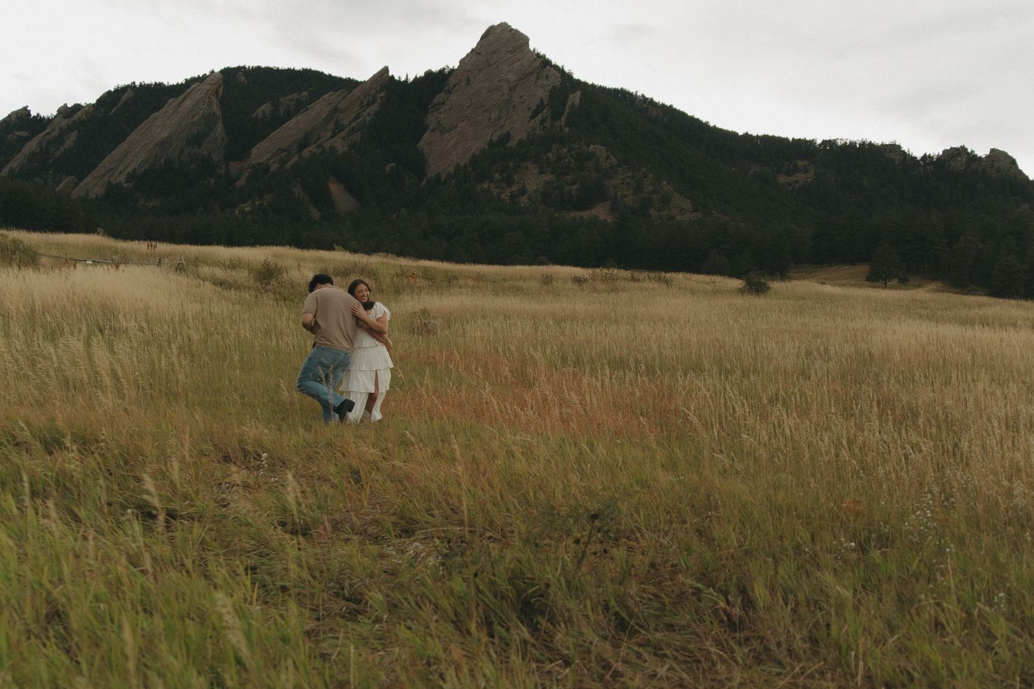 Couple embracing in golden meadow with dramatic Flatirons rock formations in background.