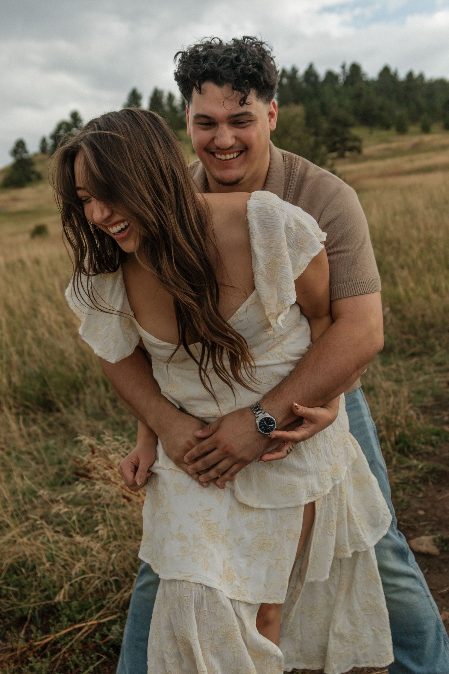 Joyful couple laughing and embracing in a golden grass field with forested mountains in the background.