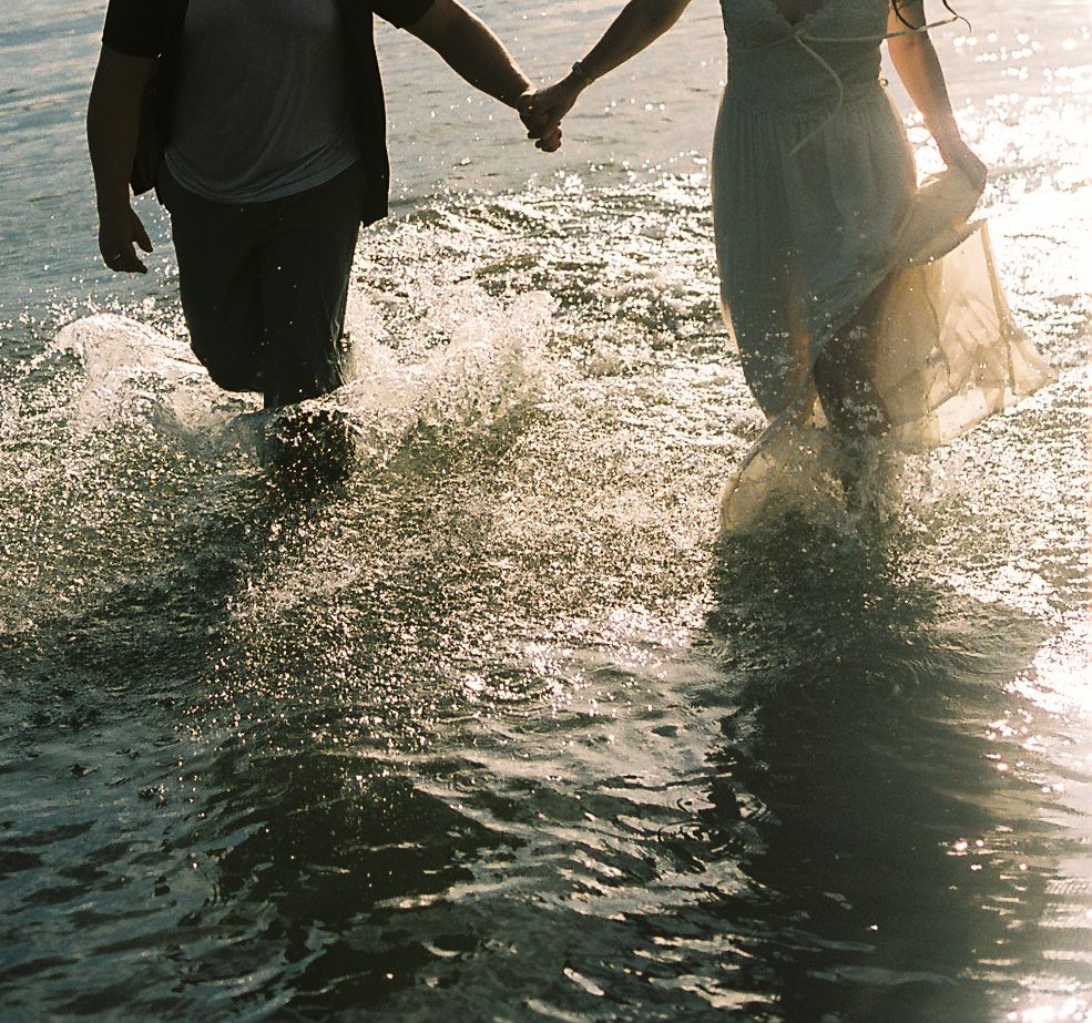 Couple holding hands and splashing through shallow lake water in golden sunlight during a playful engagement photo session.