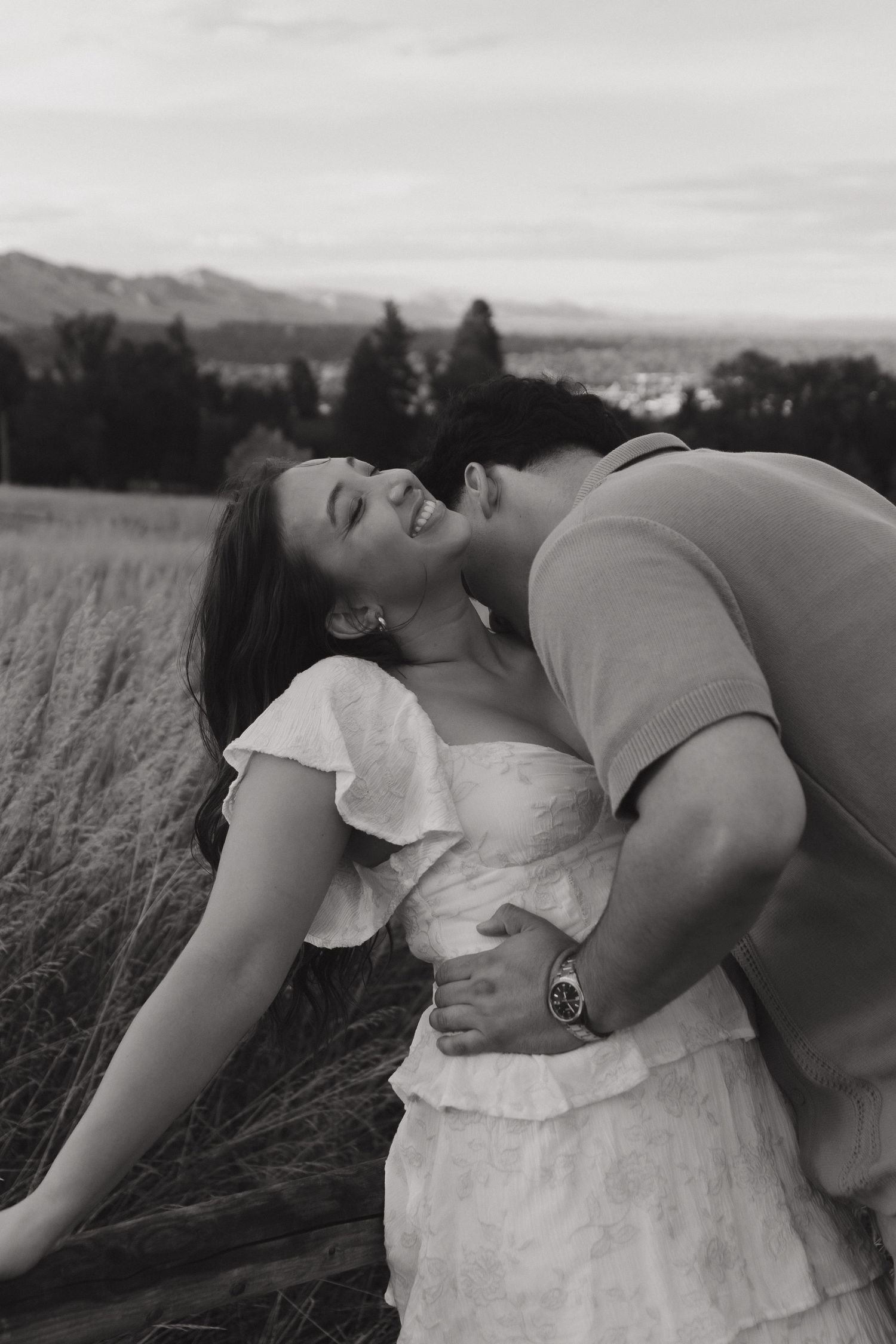 Black and white photo of man kissing woman on cheek near a rustic fence in a field.