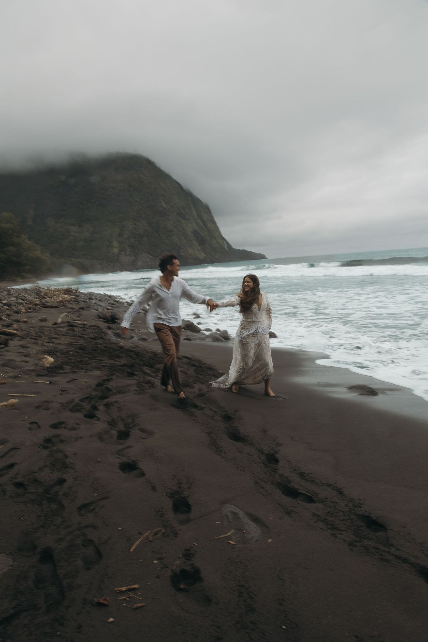 Newlyweds walk hand-in-hand along black sand beach with misty mountain cliffs and ocean waves.