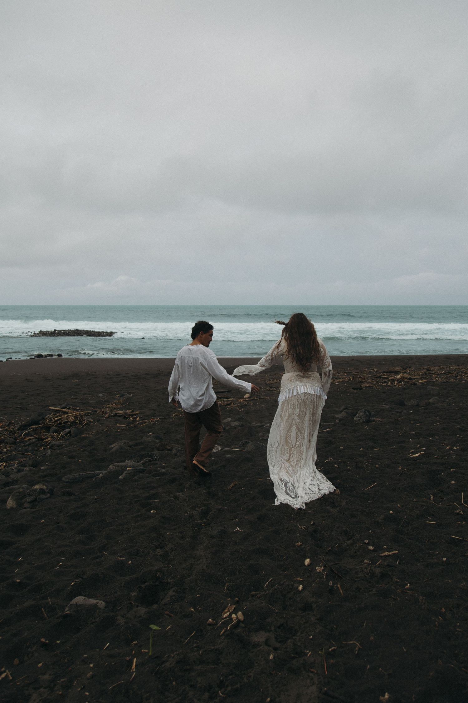 Bride in lace gown and groom holding hands walking on dark volcanic beach with moody ocean waves.