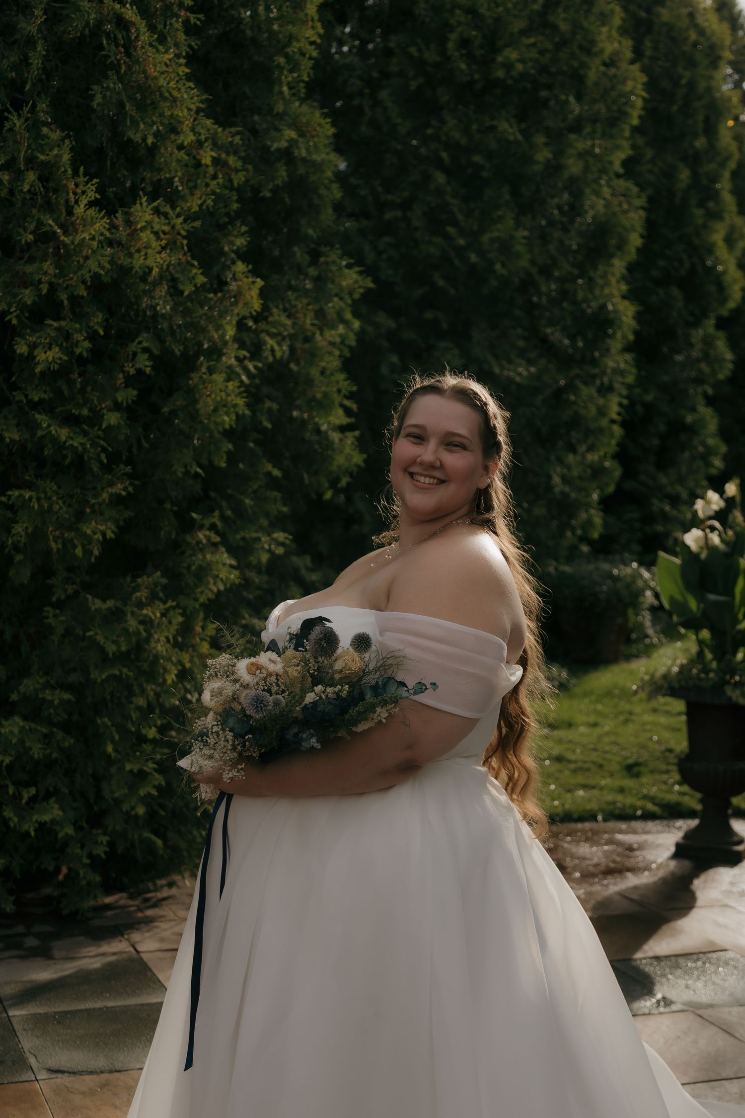 Smiling bride in off-shoulder white gown holding a floral bouquet, posing outdoors among lush green hedges.