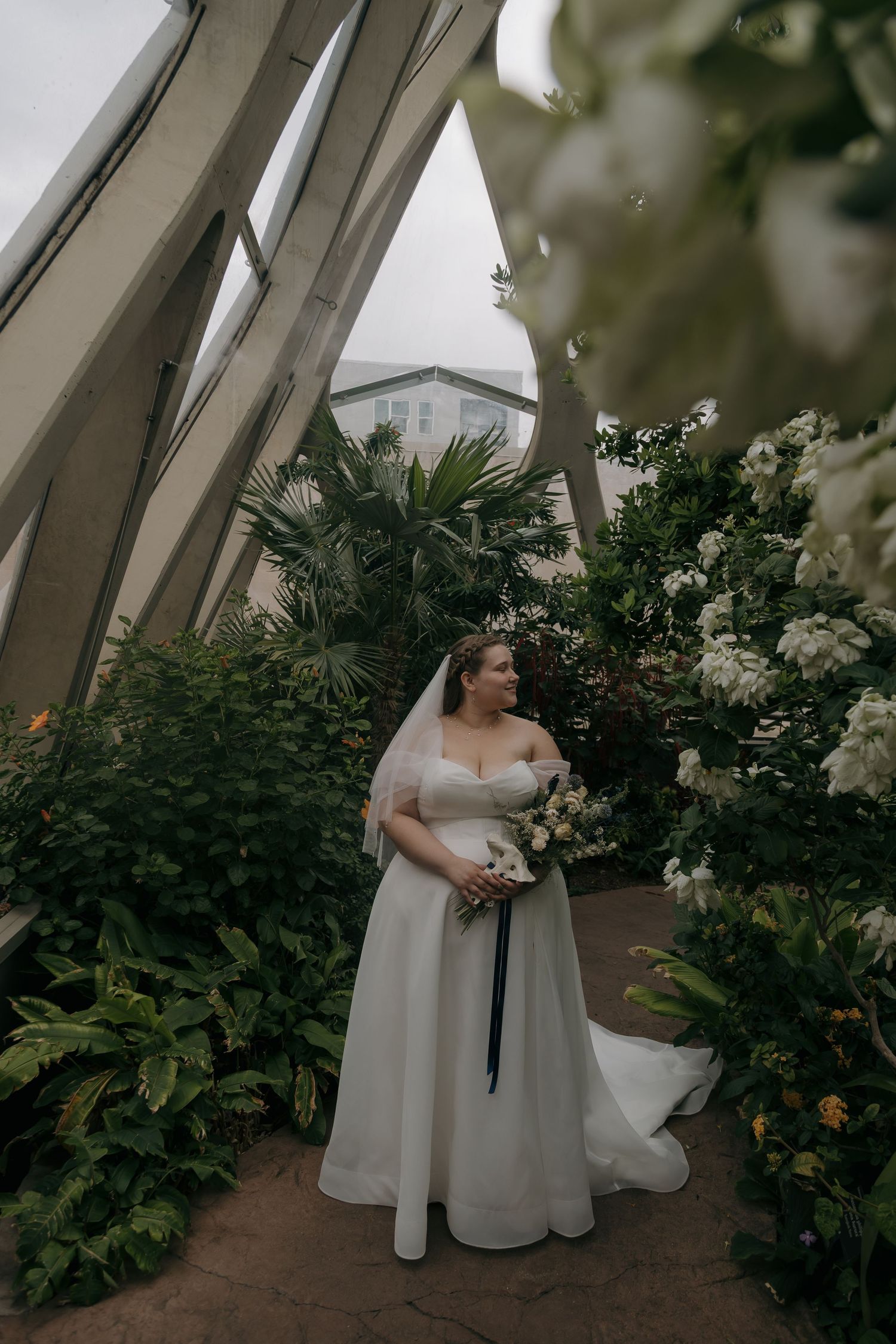Bride in white gown and veil holds bouquet surrounded by lush greenery and white flowers in a greenhouse venue.