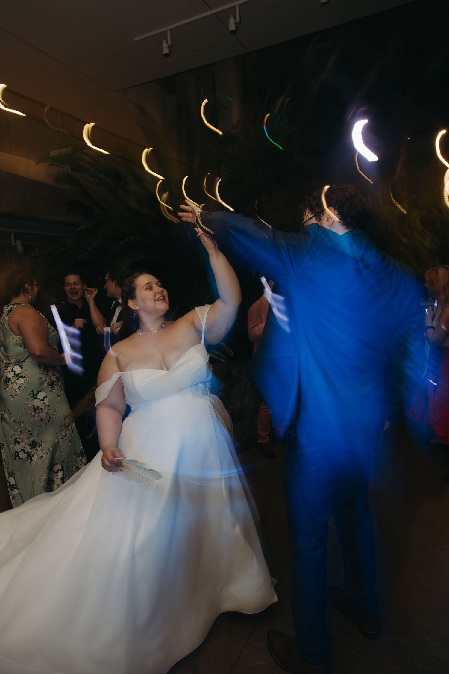 Bride in white off-shoulder gown dances with groom in blue suit under colorful reception lights.