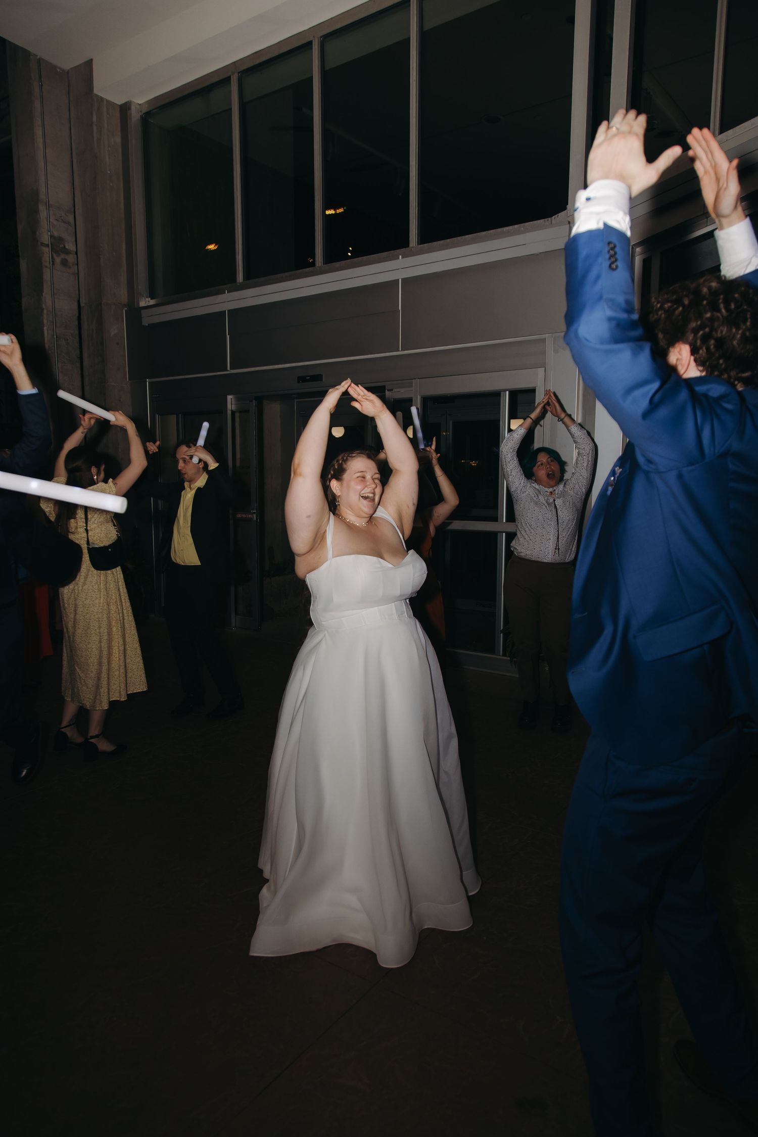 Bride in white gown dances joyfully with hands raised among celebrating wedding guests on the reception dance floor.