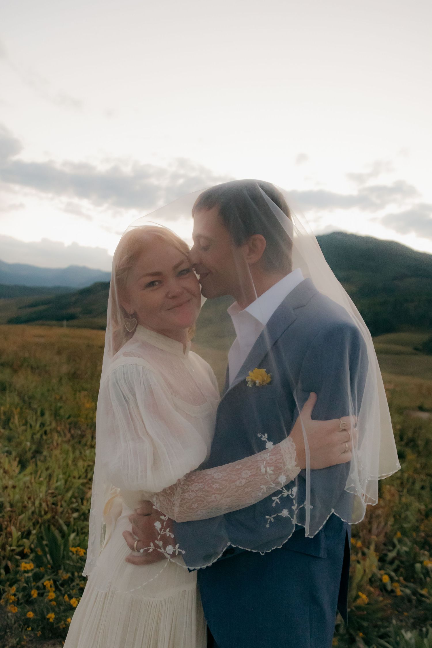 Wedding couple embracing under a veil with mountain meadow backdrop at golden hour sunset.